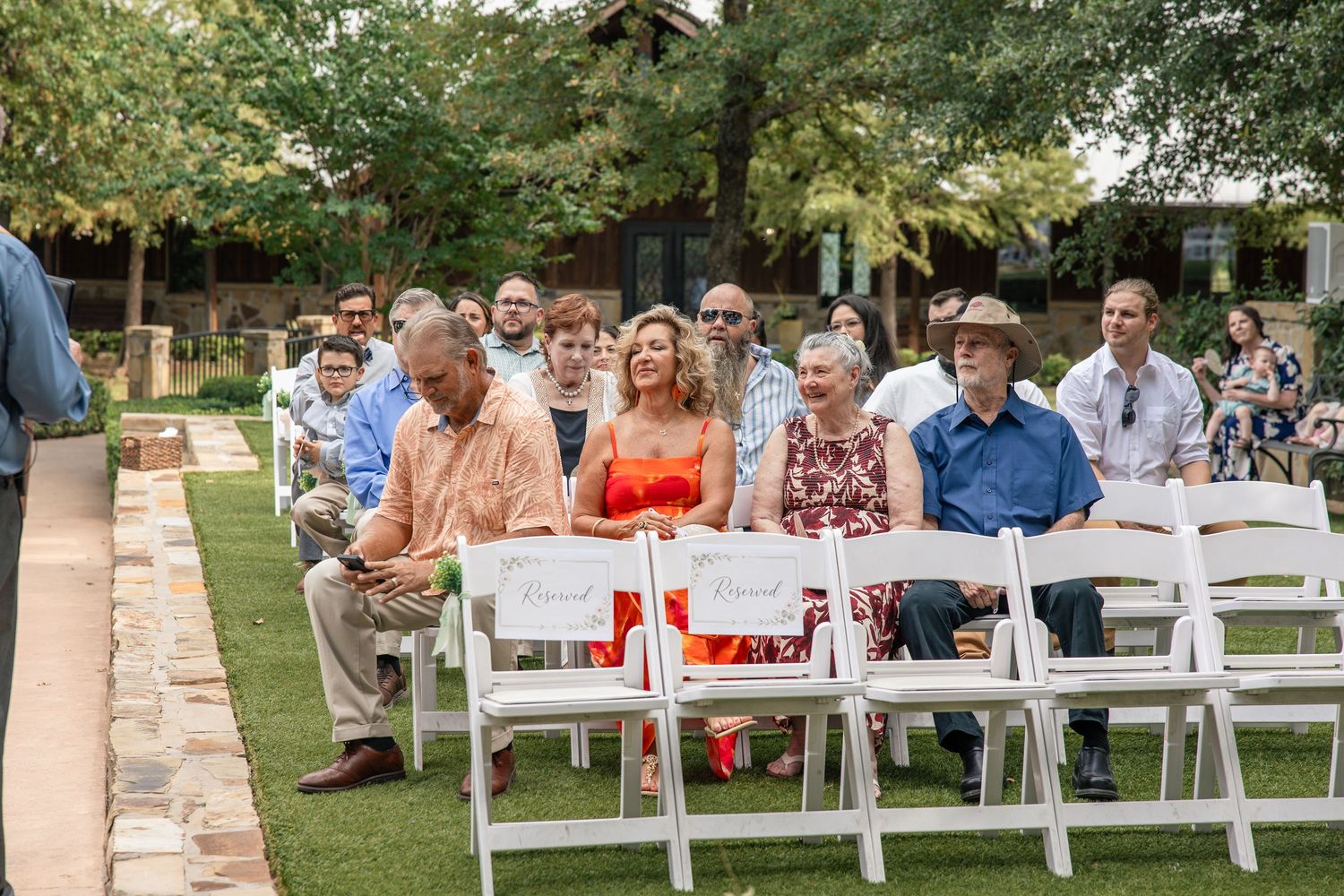 Wedding guests in summer attire sit on white folding chairs on a grassy lawn, watching an outdoor ceremony.