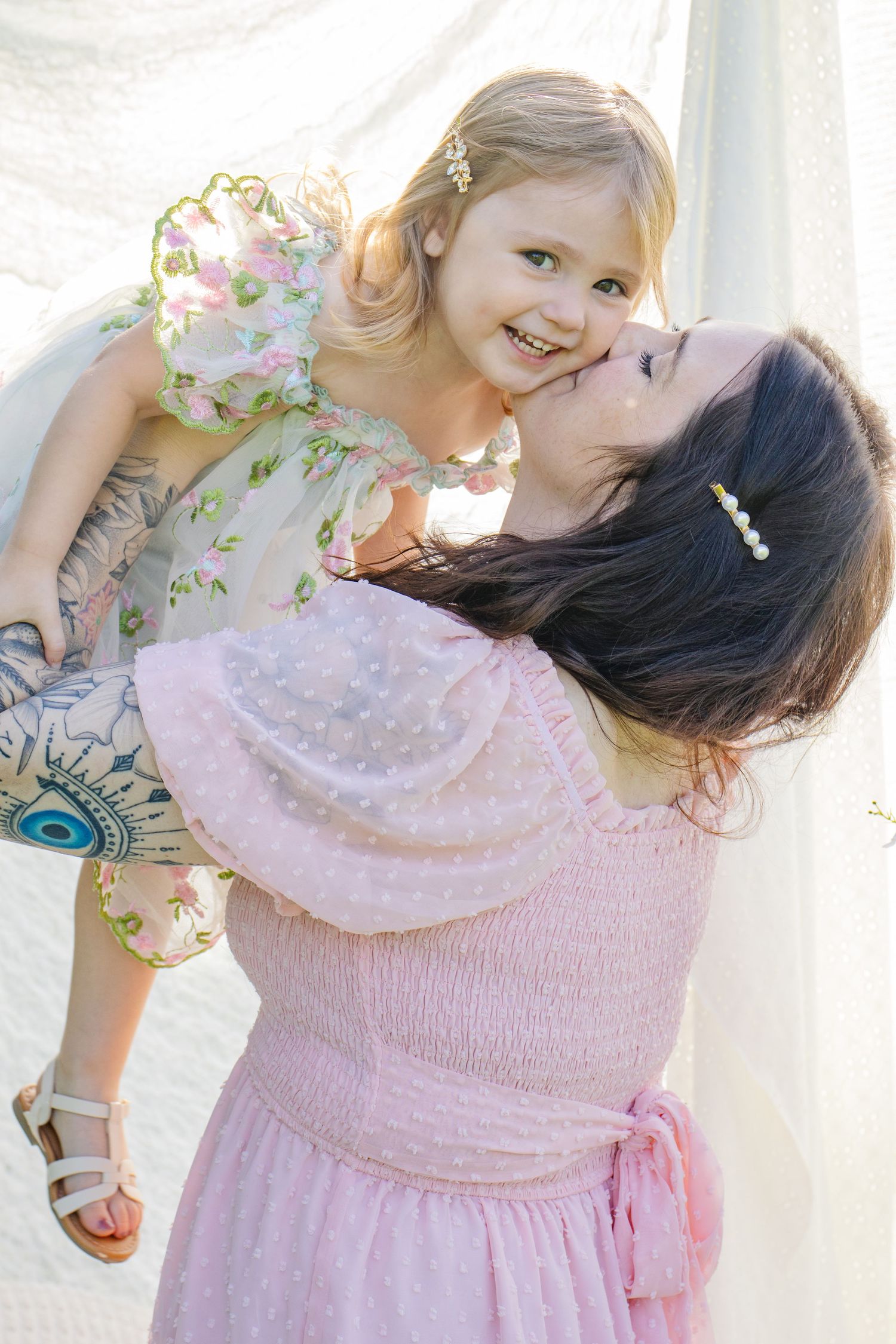 A mother in a pink dress lovingly holds her smiling toddler daughter in a floral dress outdoors.