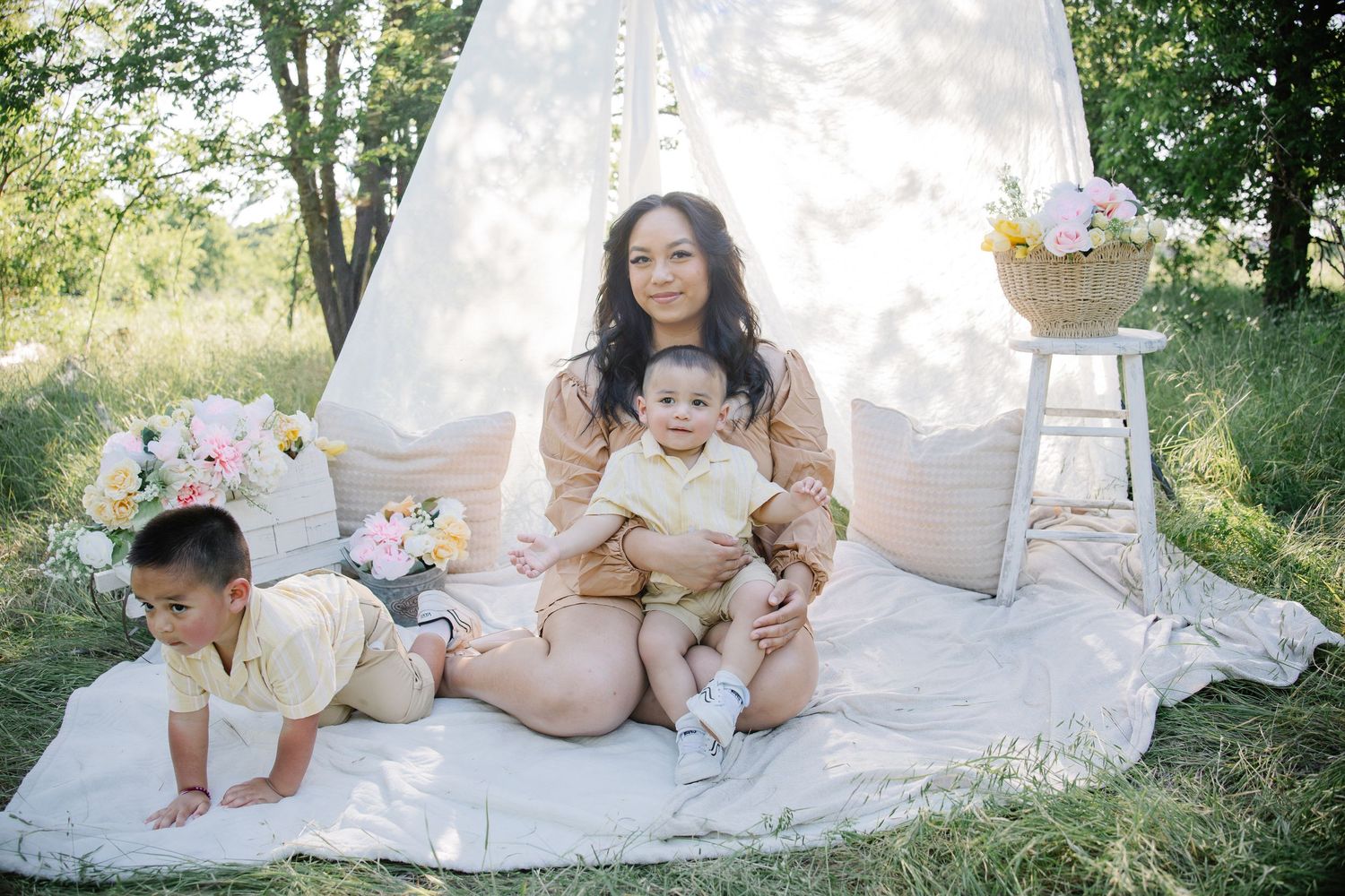 A mother sits outdoors with two young children near a white teepee tent, surrounded by floral arrangements on a blanket.