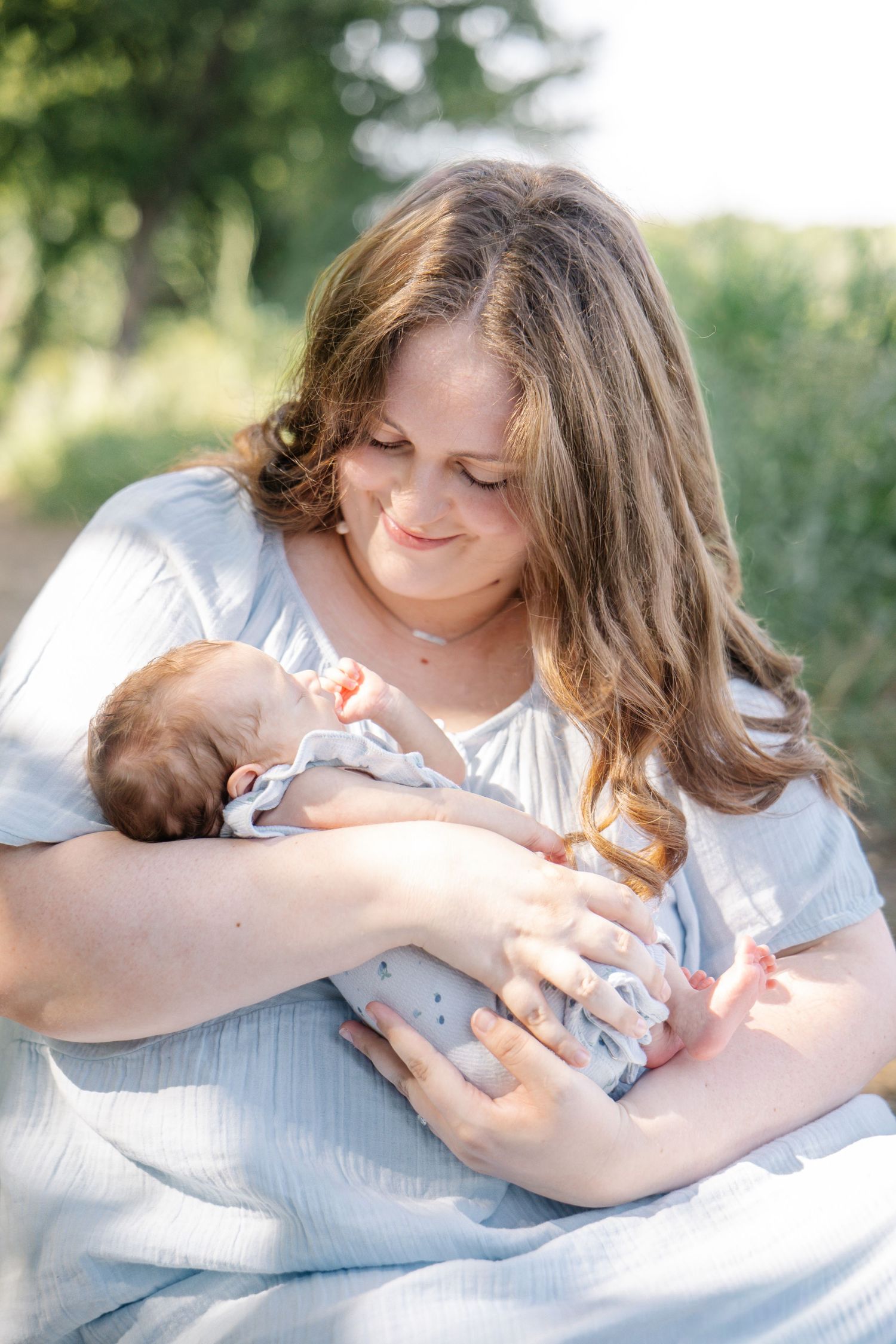 A young mother in a light blue dress tenderly holds her newborn baby outdoors in soft natural light.