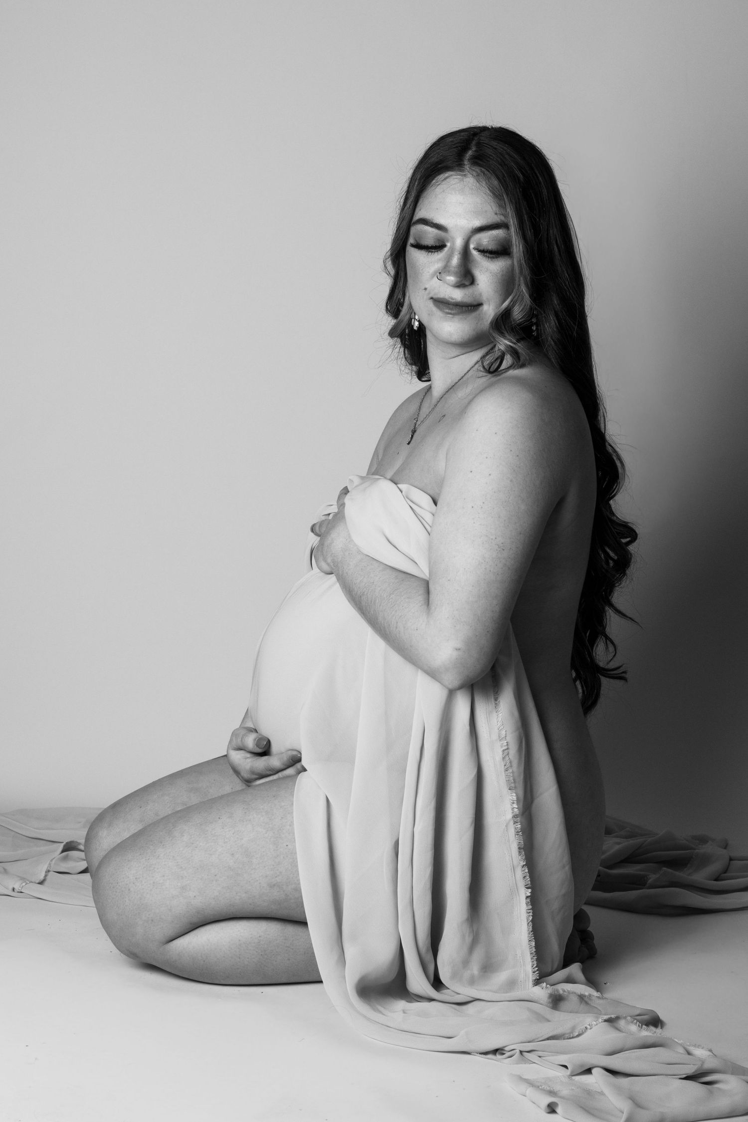 Black and white maternity photo of a pregnant woman kneeling, cradling her baby bump in a flowing draped fabric.