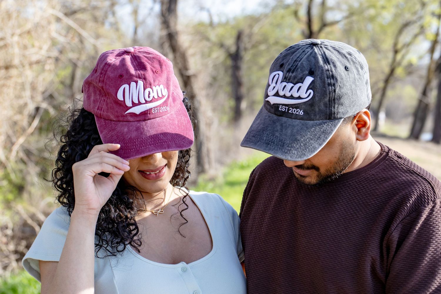Expecting couple wearing matching Mom and Dad baseball caps smiling outdoors among trees in natural light.
