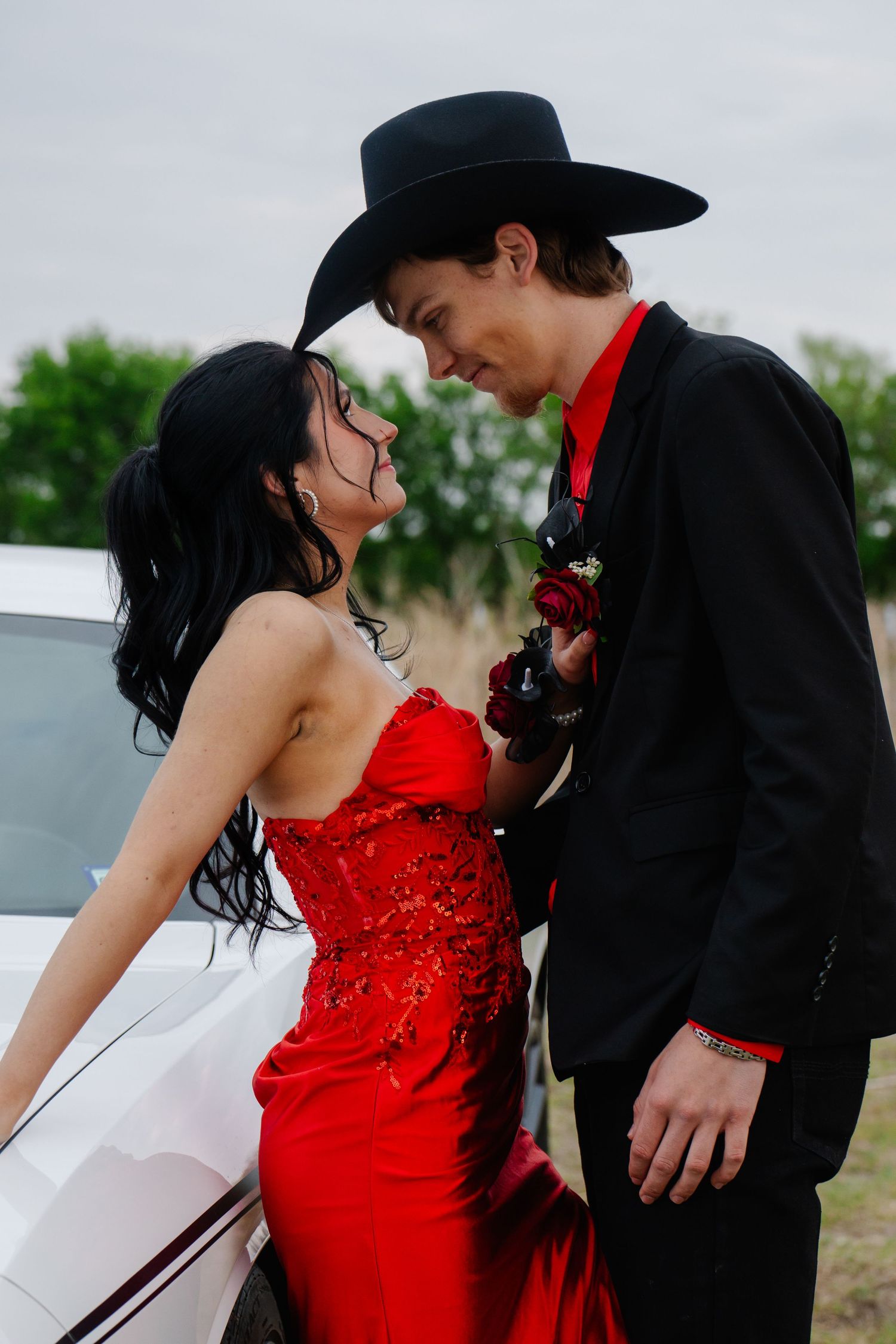 Prom couple leaning against white car, girl in red gown, boy in black suit with cowboy hat.
