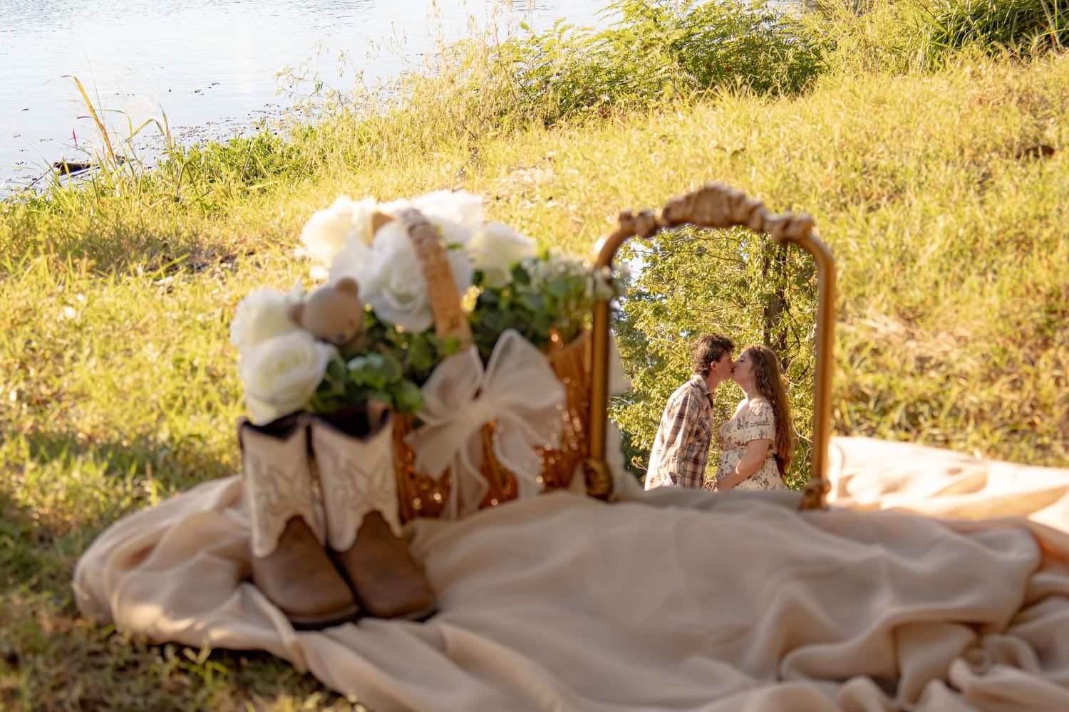 Vintage mirror reflects a kissing couple beside cowboy boots and a white rose bouquet in a rustic outdoor flat lay.
