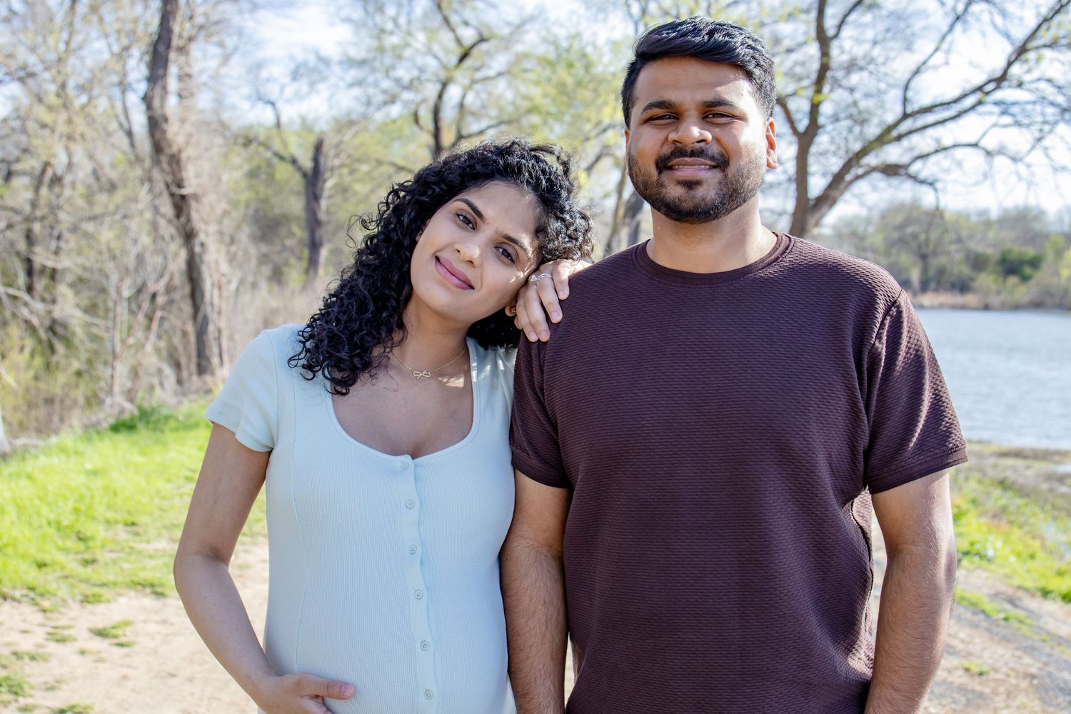 Smiling South Asian couple posing together near a lake surrounded by lush green trees outdoors.