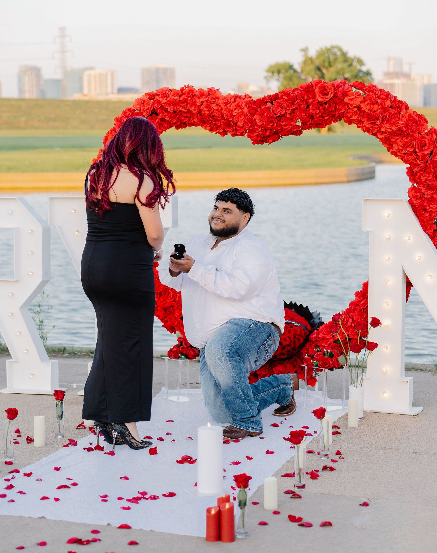 Man proposing under red rose heart arch with candles and rose petals by waterfront at sunset.