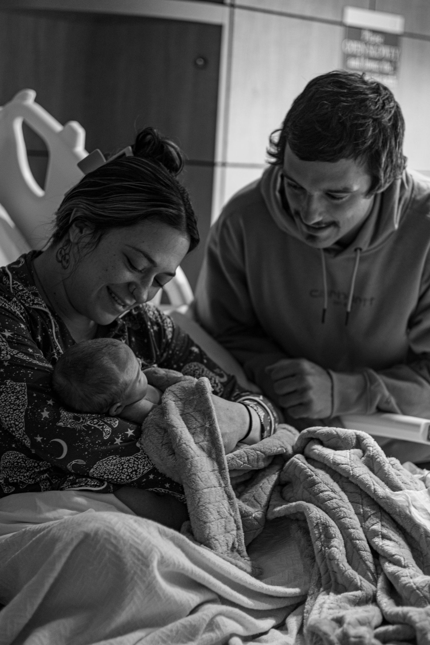 New parents smiling together holding newborn baby in hospital room, black and white.