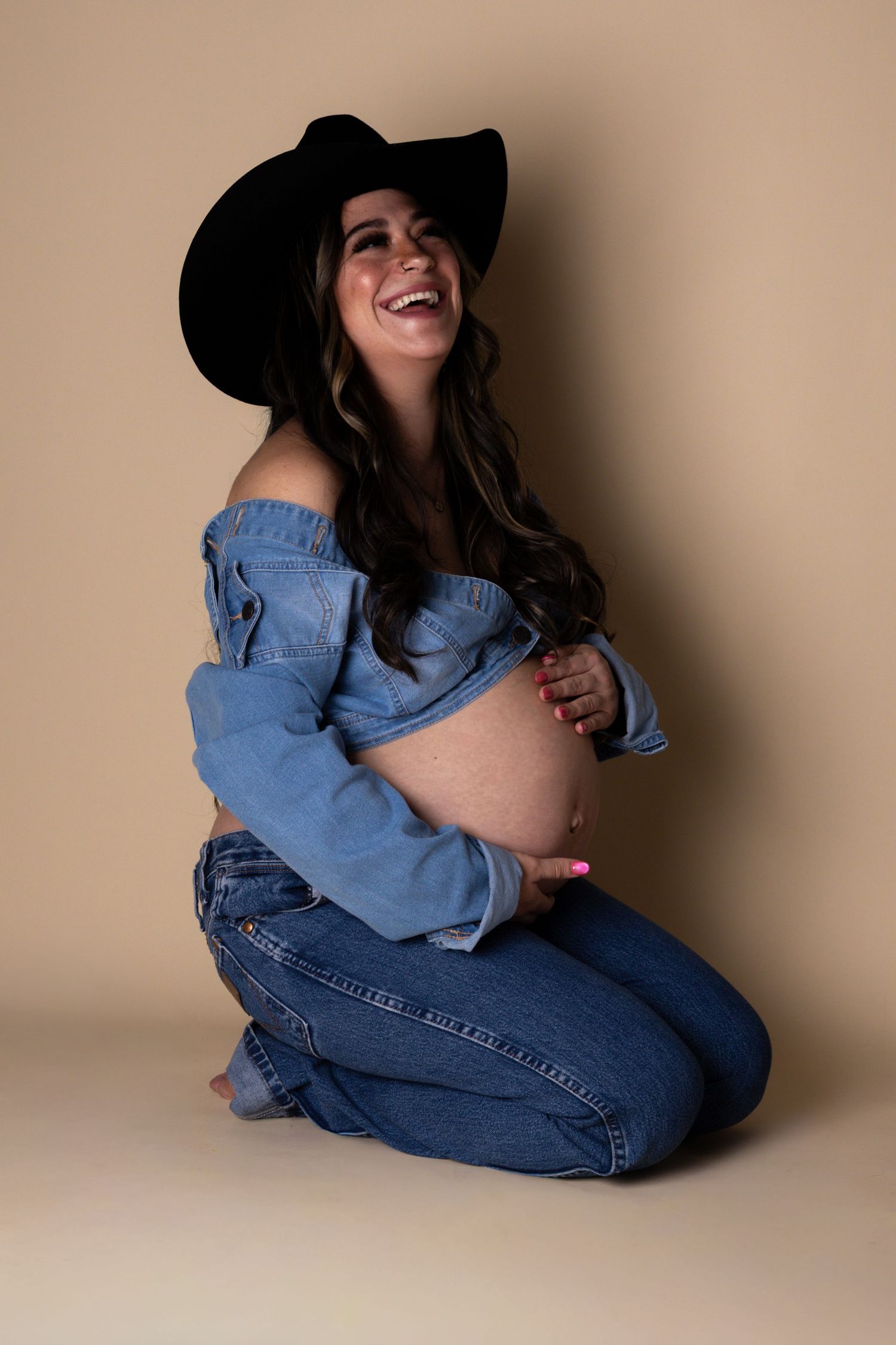 Laughing pregnant woman in denim off-shoulder top and black cowboy hat kneeling on studio floor.