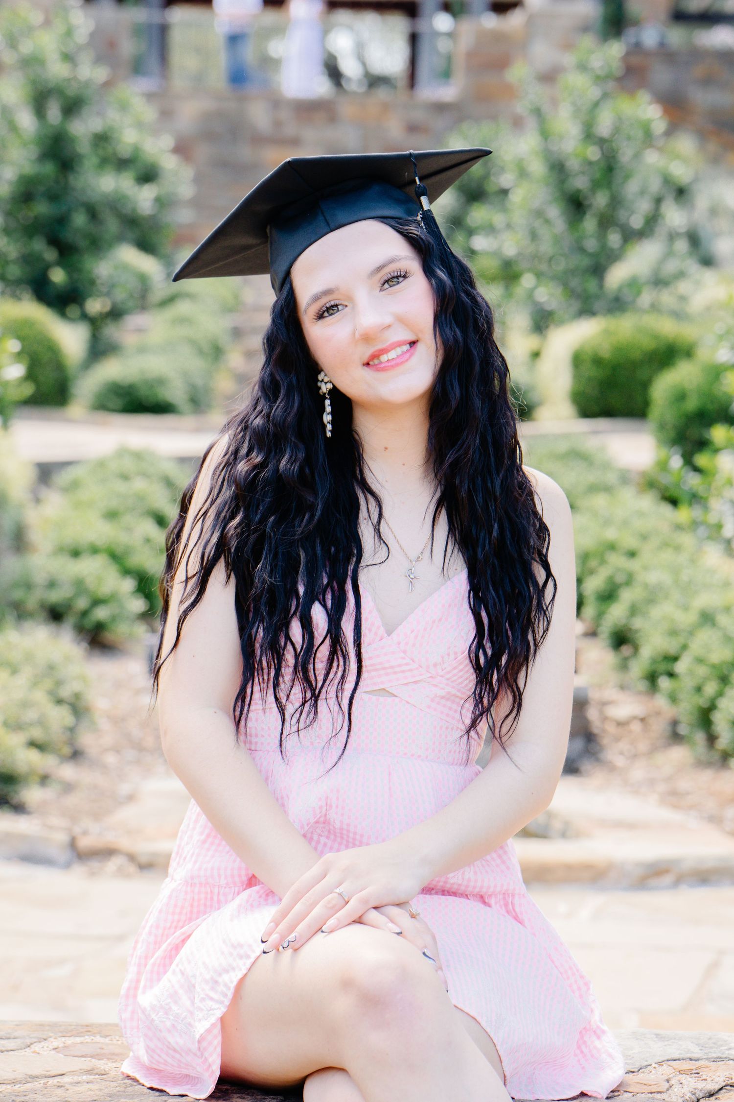 Beautiful graduate smiling in black cap and pink dress seated in lush garden for senior portraits.