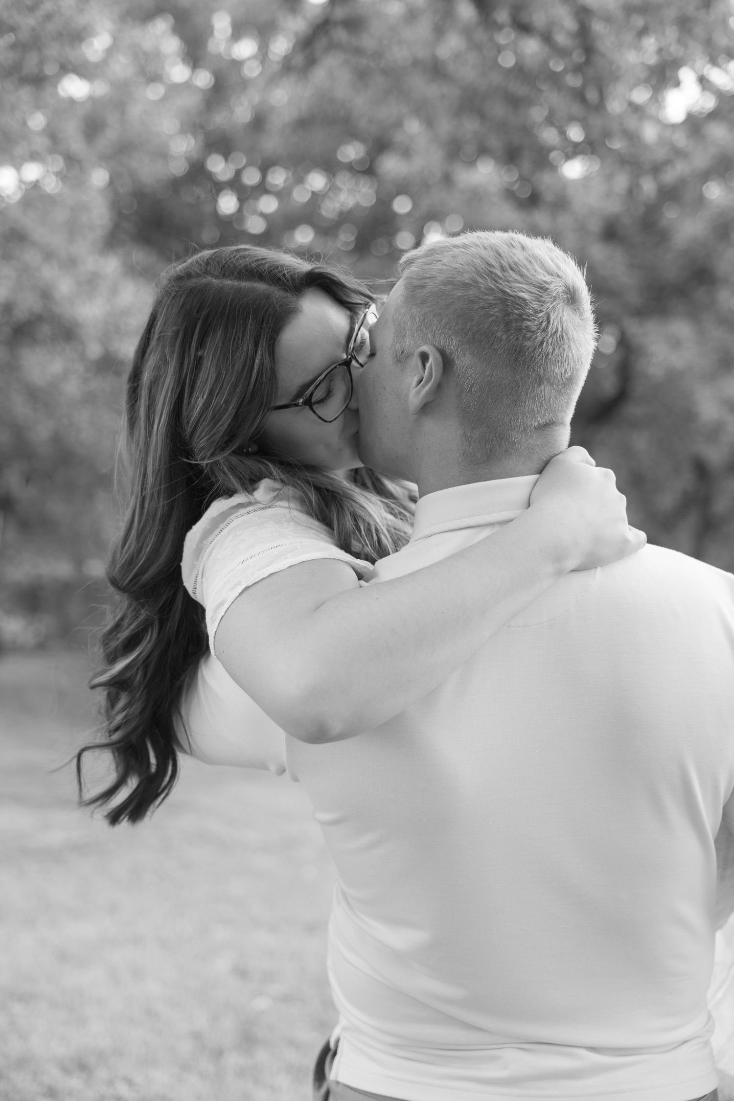Black and white photo of engaged couple sharing a kiss in an outdoor park setting.