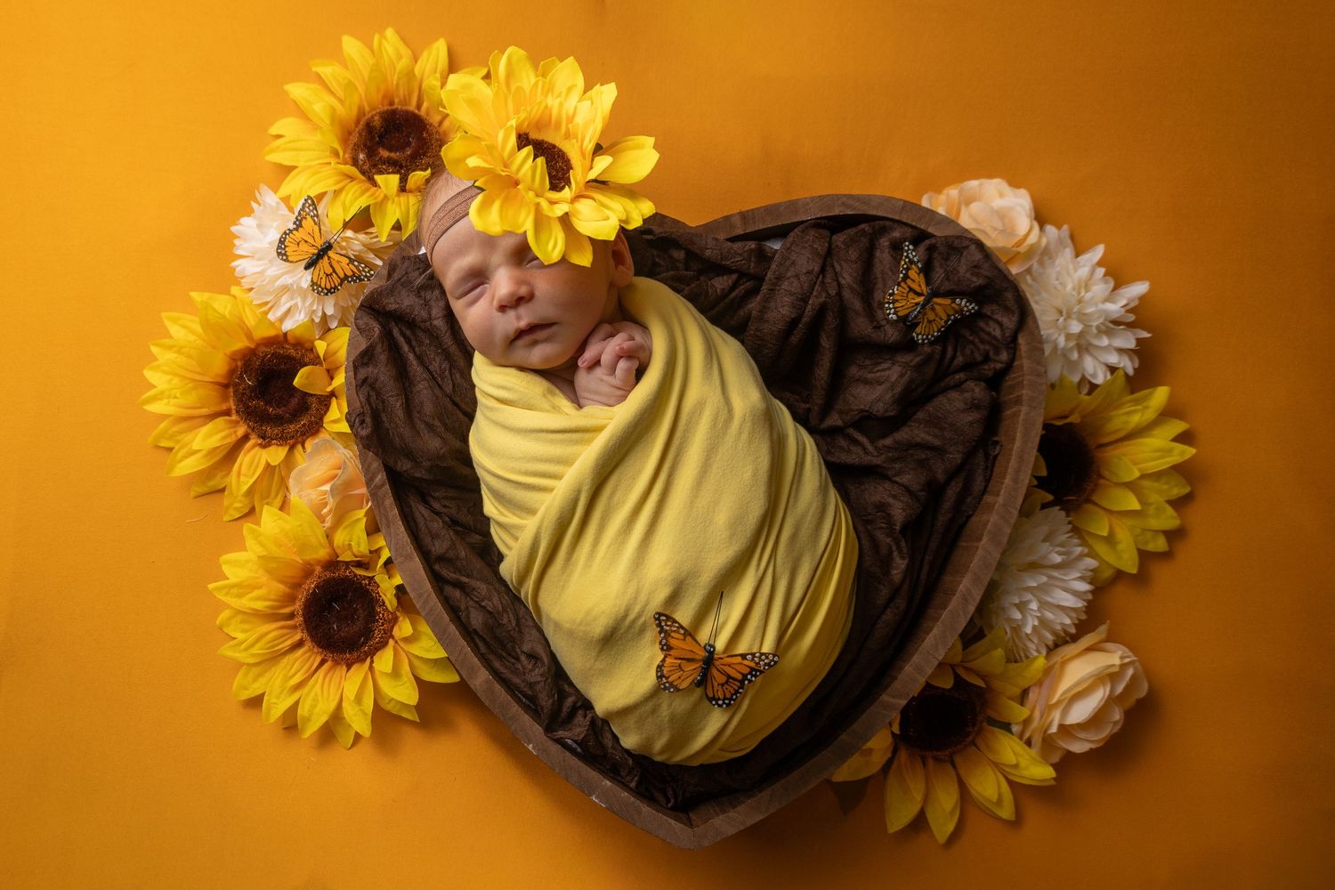 Newborn baby swaddled in yellow wrap sleeping in heart-shaped wooden bowl surrounded by sunflowers on golden background.