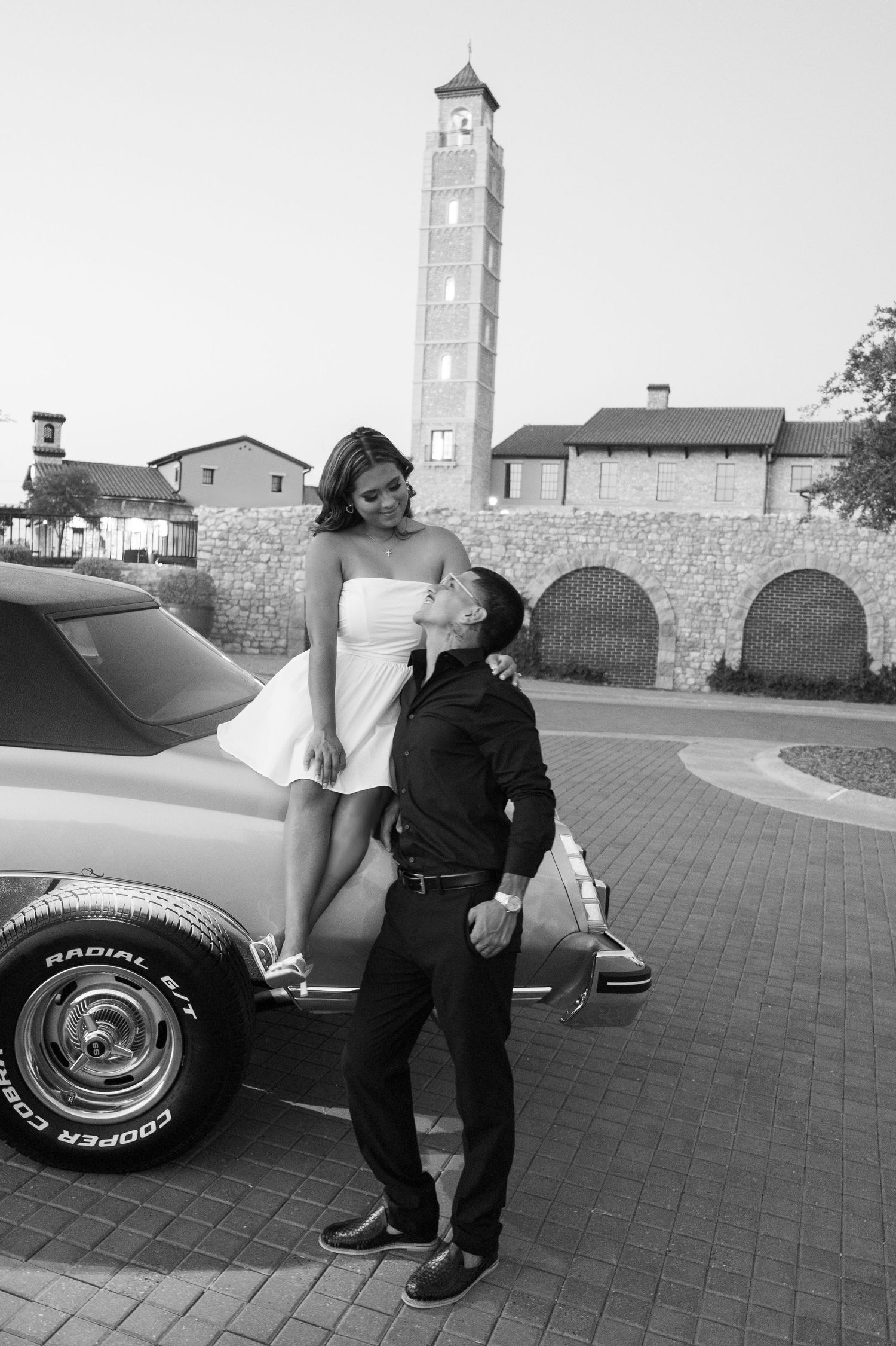 Couple posing by a classic car with a stone tower backdrop in black and white.