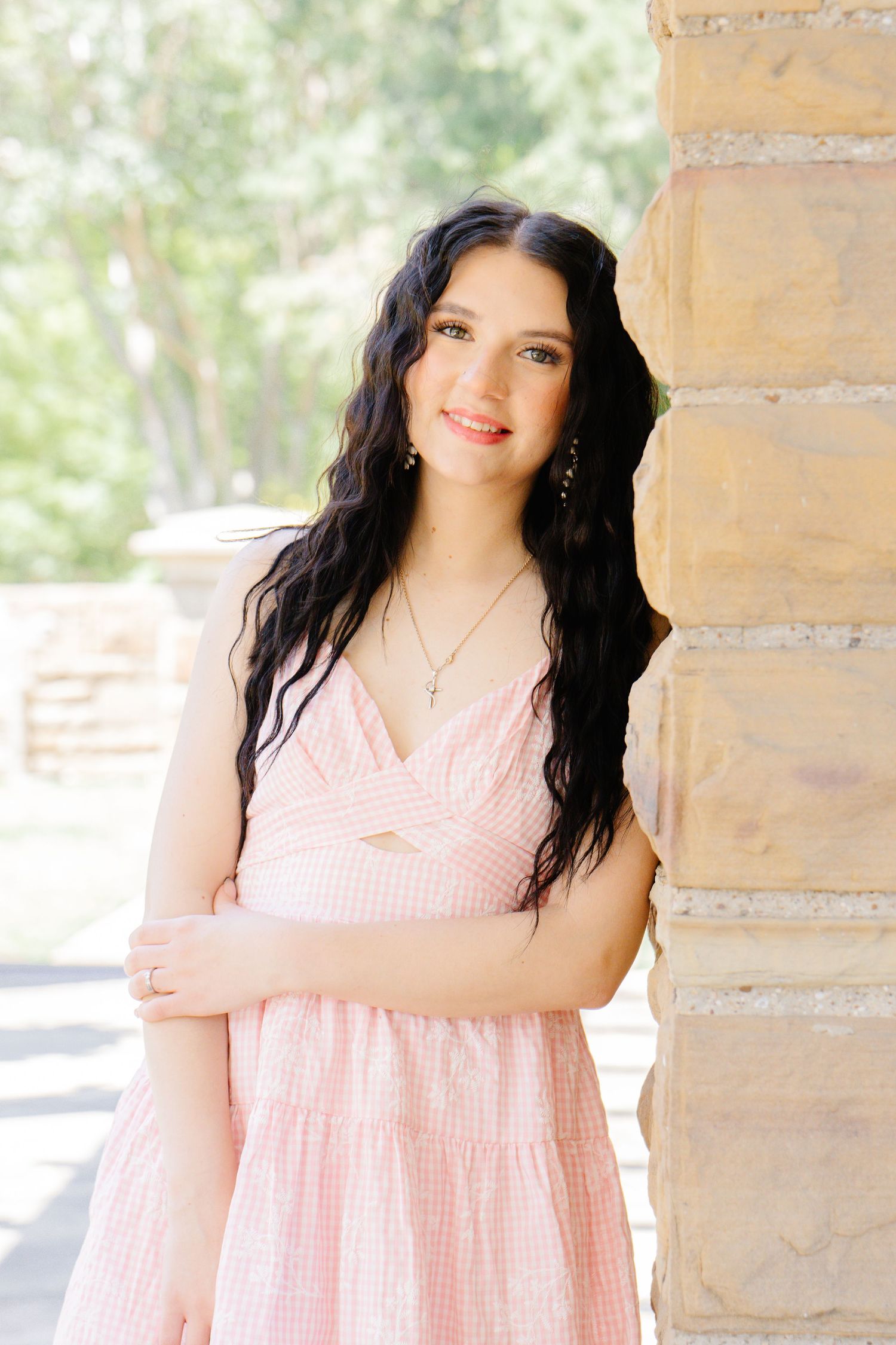 Young woman in pink dress leaning against stone pillar outdoors, wearing cross necklace and dangling earrings.