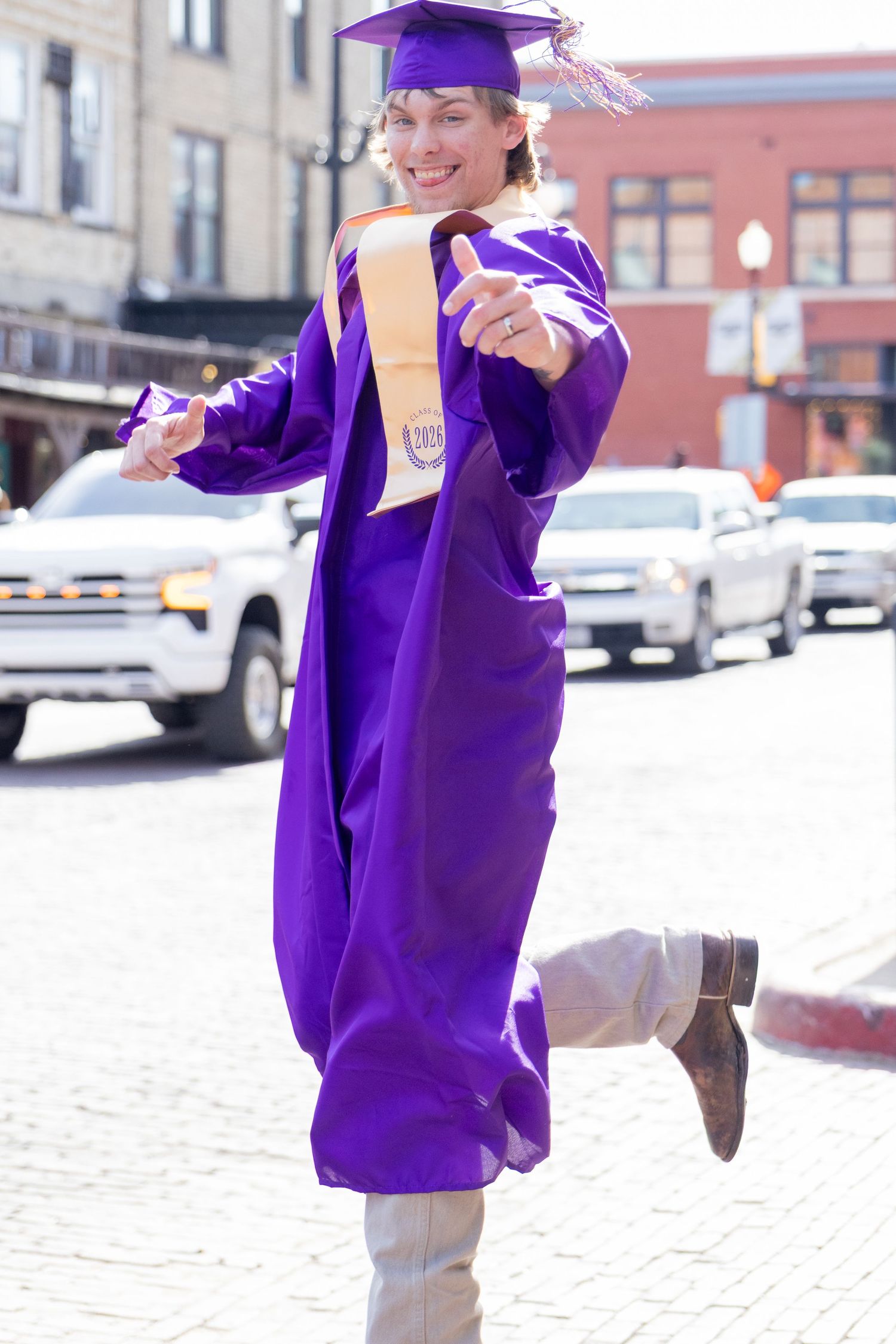 Smiling graduate in purple cap and gown holding diploma outdoors on sunny street.