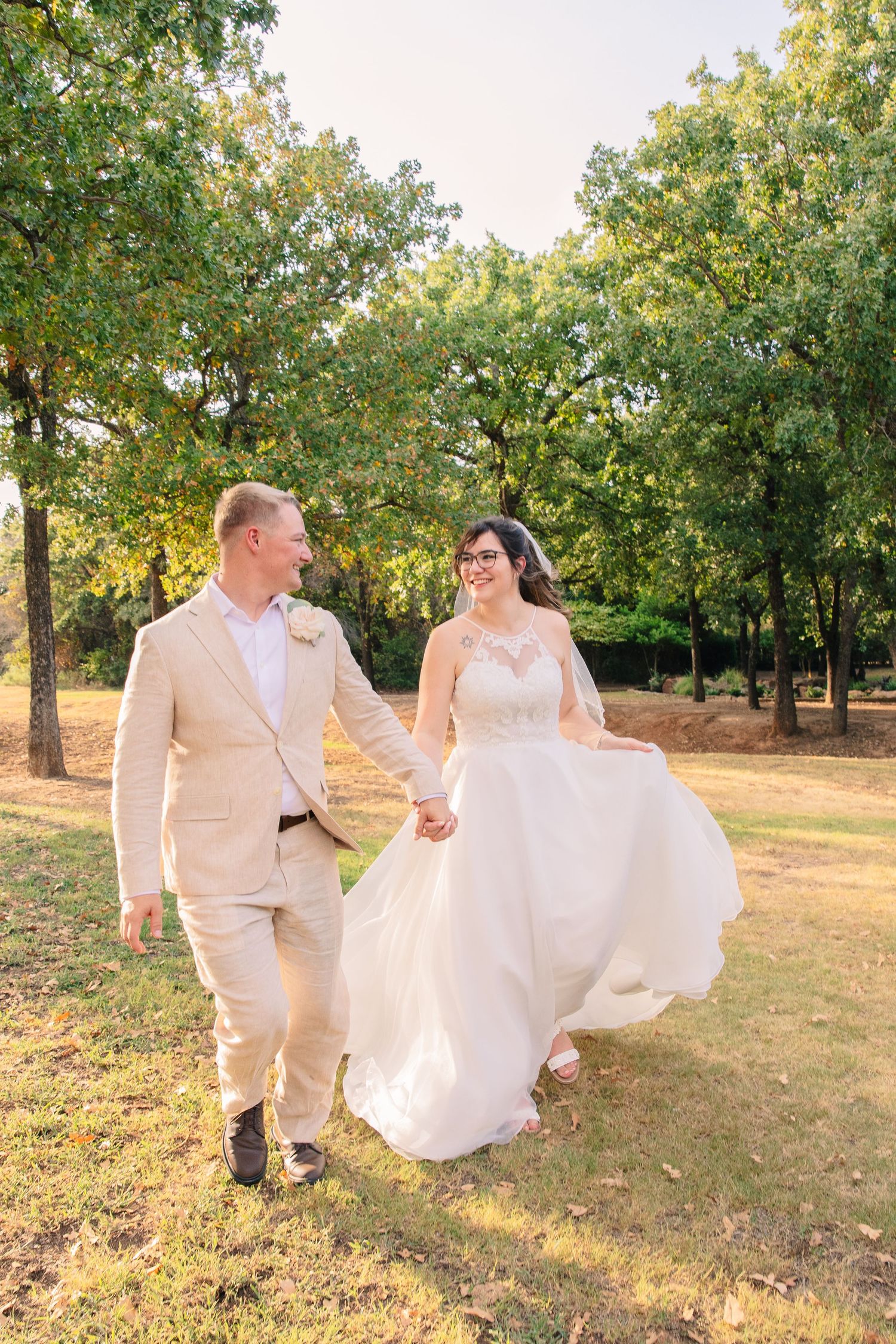 Smiling bride and groom hold hands and walk through a sunny park, her flowing white wedding dress swaying beneath tall green trees.