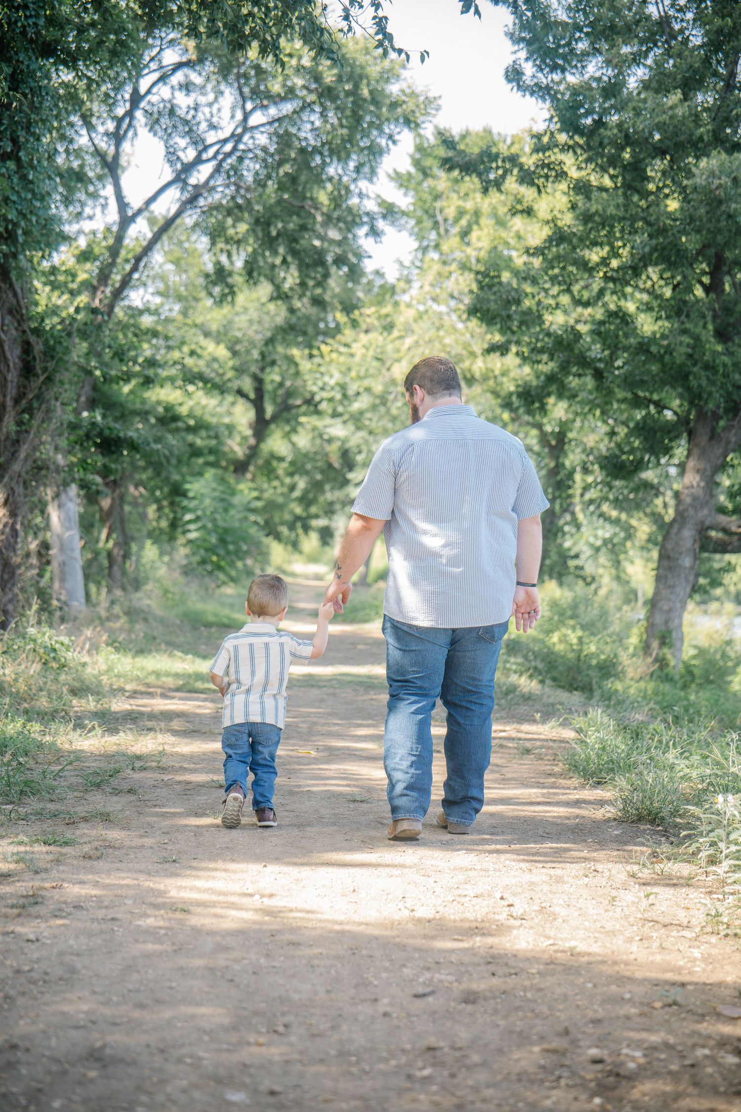 Father and young son walking hand in hand down a peaceful dirt path surrounded by lush green trees.