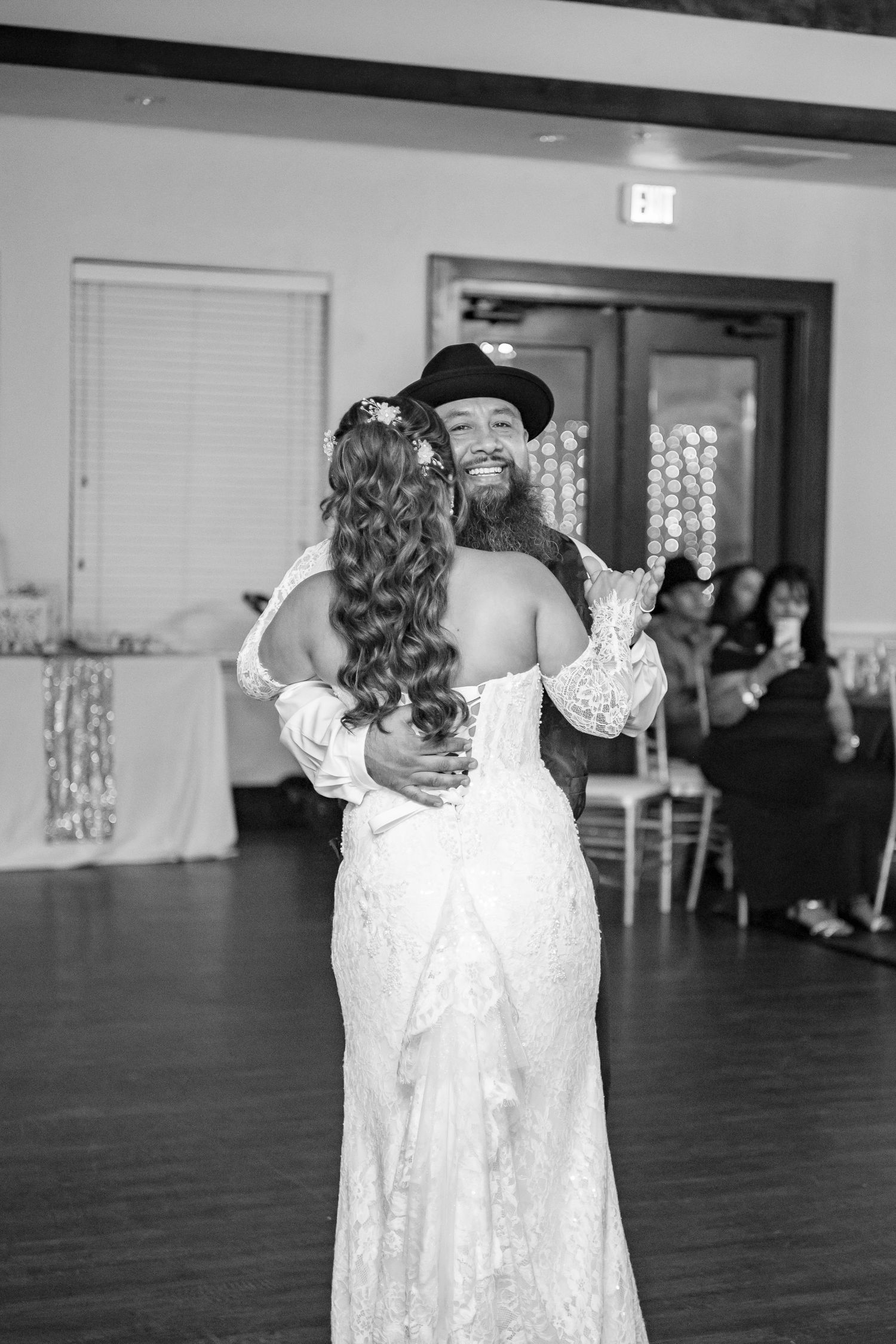 Black and white photo of a smiling groom in a hat sharing a first dance with his bride in a lace wedding gown.