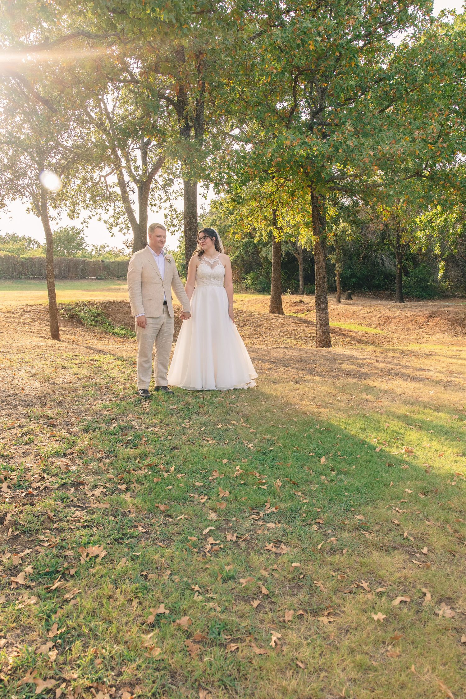 Bride in a white gown and groom in a tan suit hold hands in a sunlit, tree-lined grassy field at their outdoor wedding.