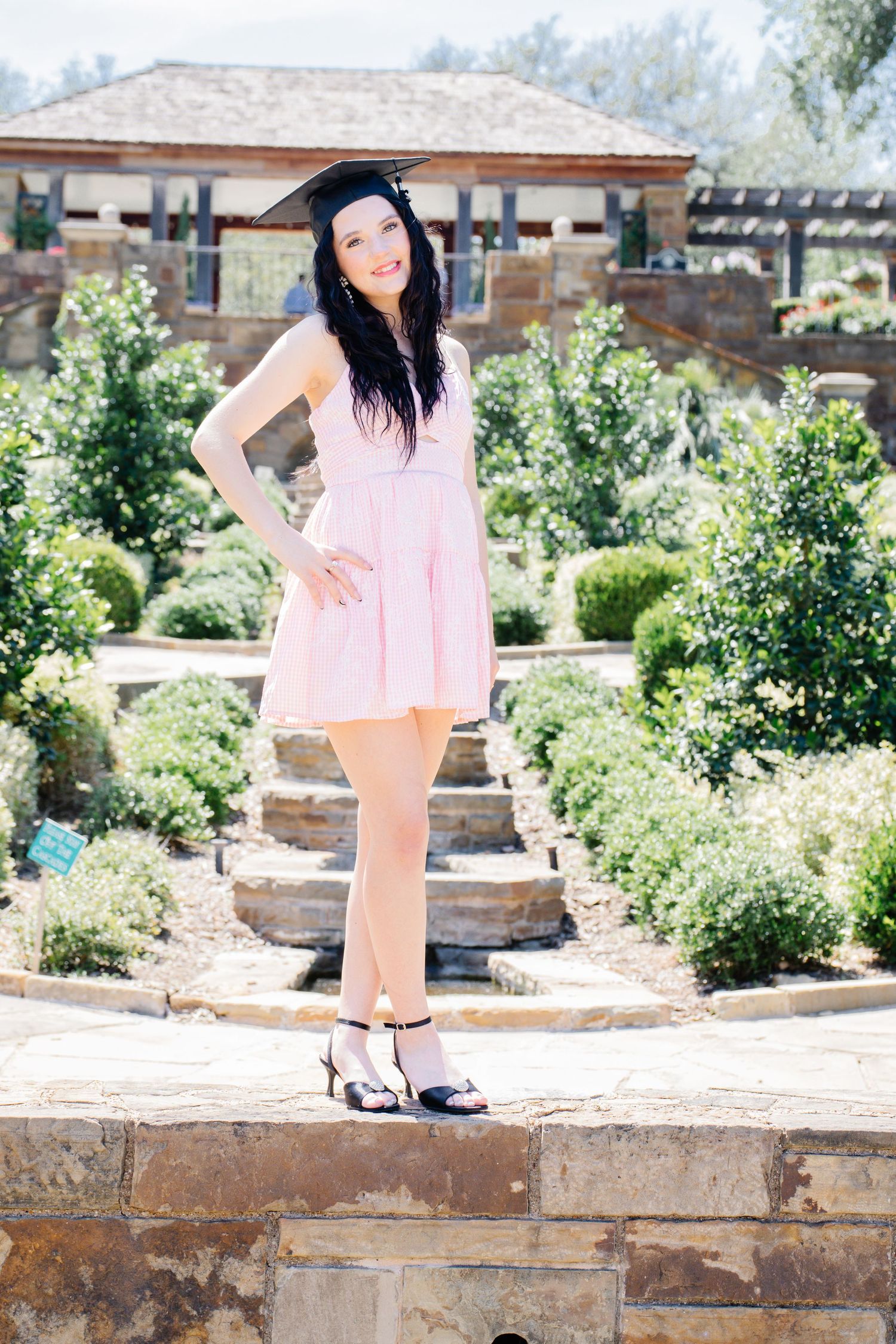 A young woman in a pink dress and graduation cap poses confidently in a beautiful garden setting.