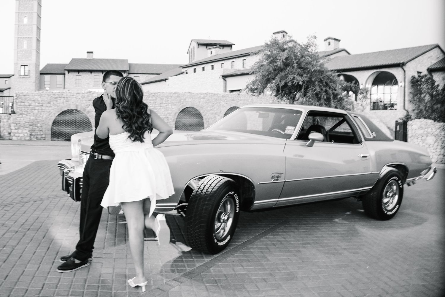Black and white photo of a couple kissing beside a classic vintage car at a wedding venue.