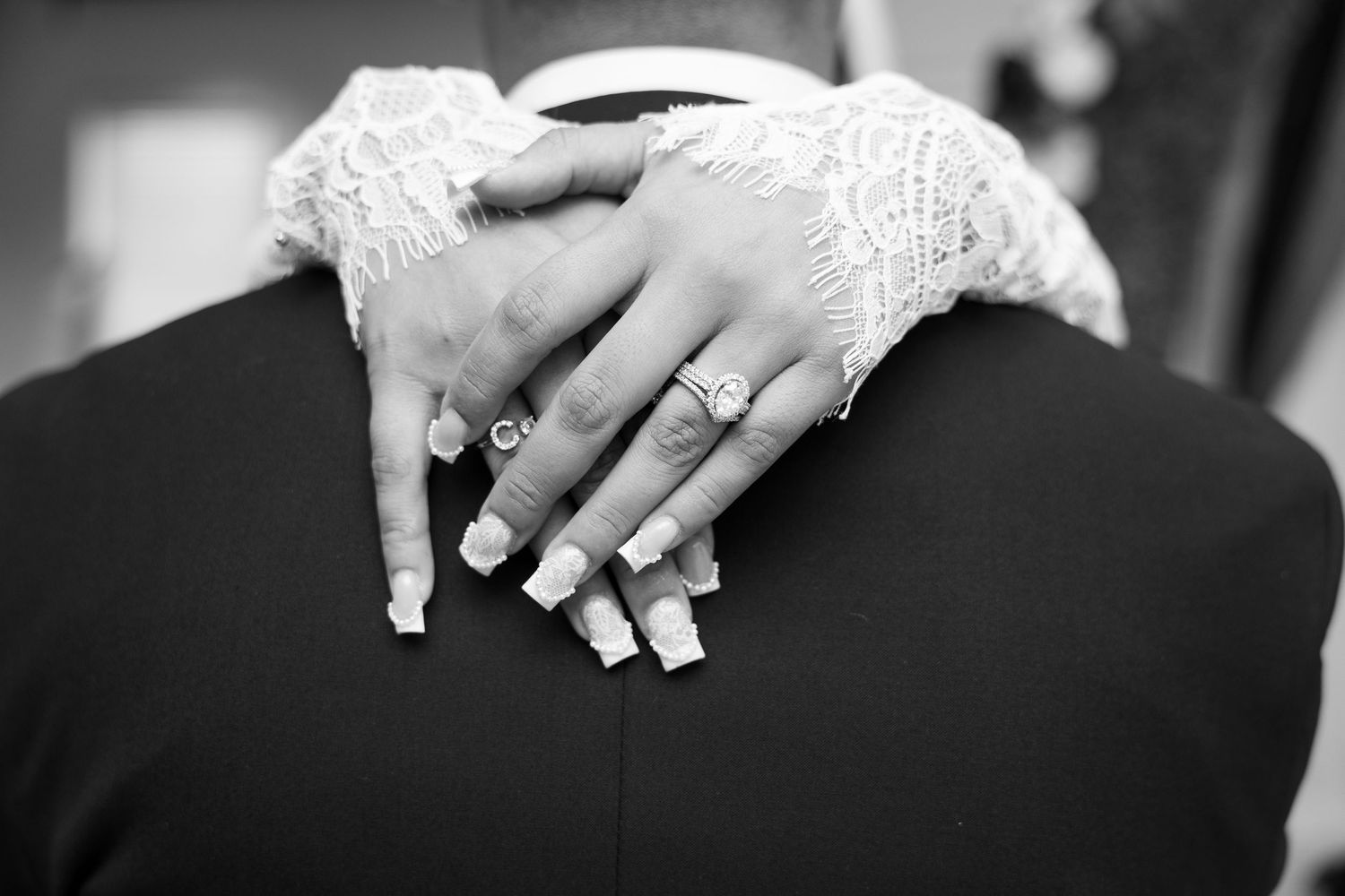 Close-up black and white photo of bride's manicured hands with rings resting on groom's suit jacket.