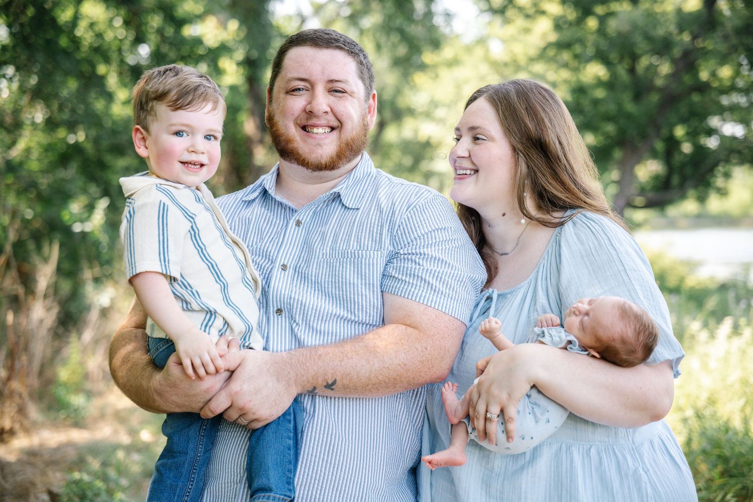 Happy family of four with toddler boy and newborn baby posing outdoors among green trees.
