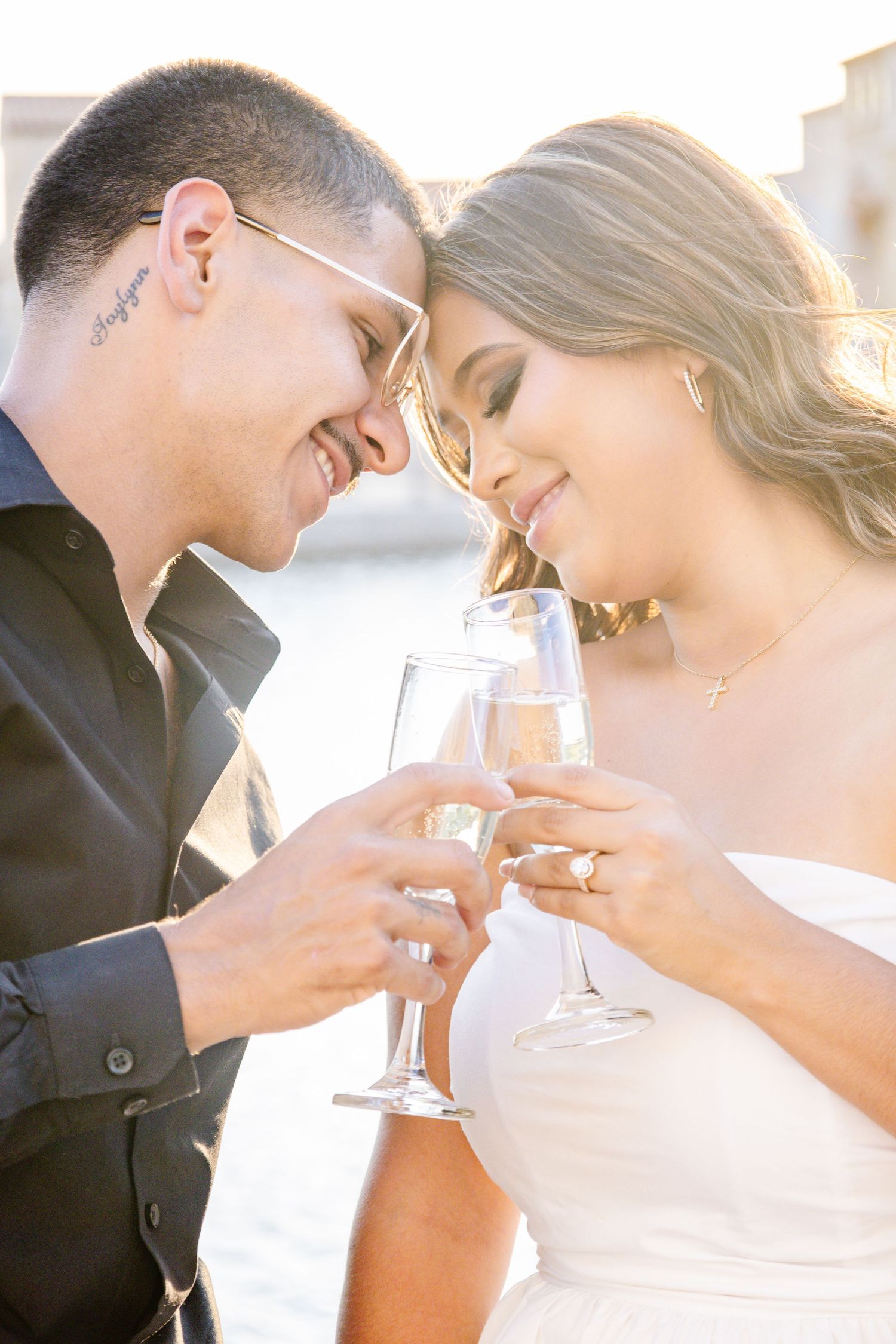 Bride and groom toasting with champagne glasses, smiling intimately in golden sunlight at waterfront wedding.