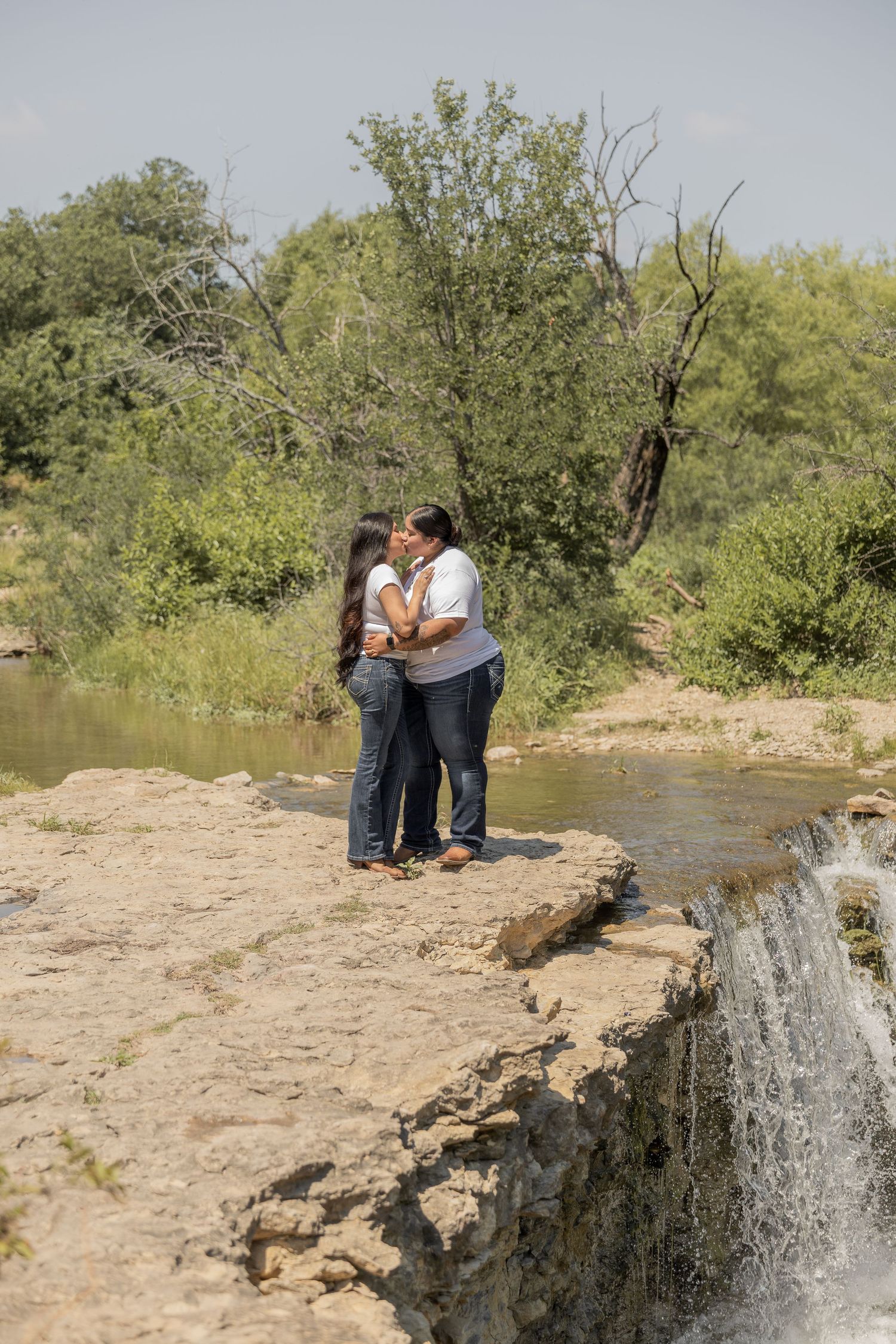 A couple shares a romantic kiss on rocky terrain beside a flowing waterfall surrounded by greenery.