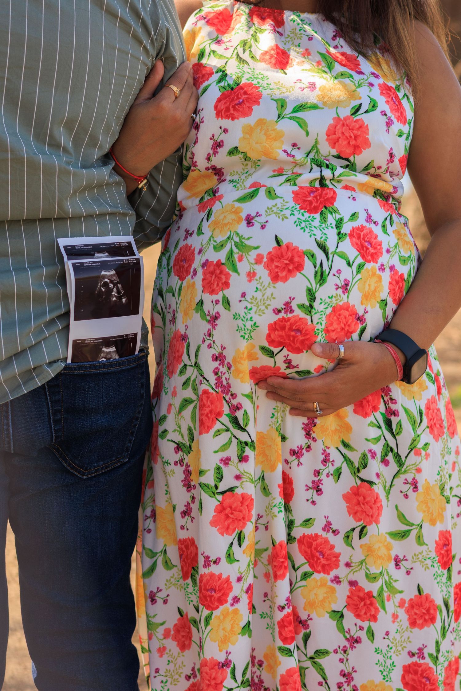 Pregnant woman in floral dress cradling baby bump beside partner holding ultrasound photos outdoors.