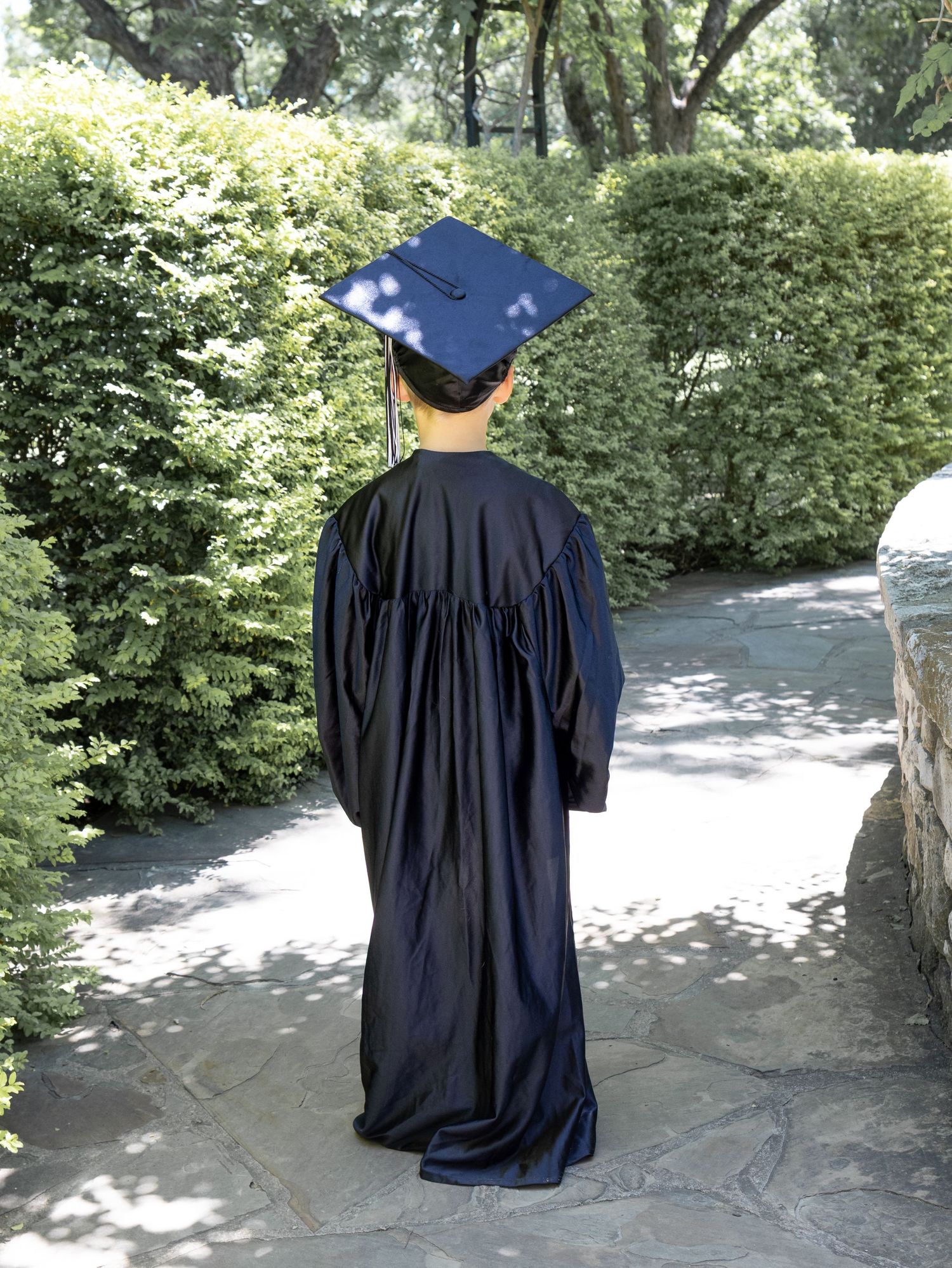 Child in navy graduation cap and gown viewed from behind on stone path surrounded by green hedges.