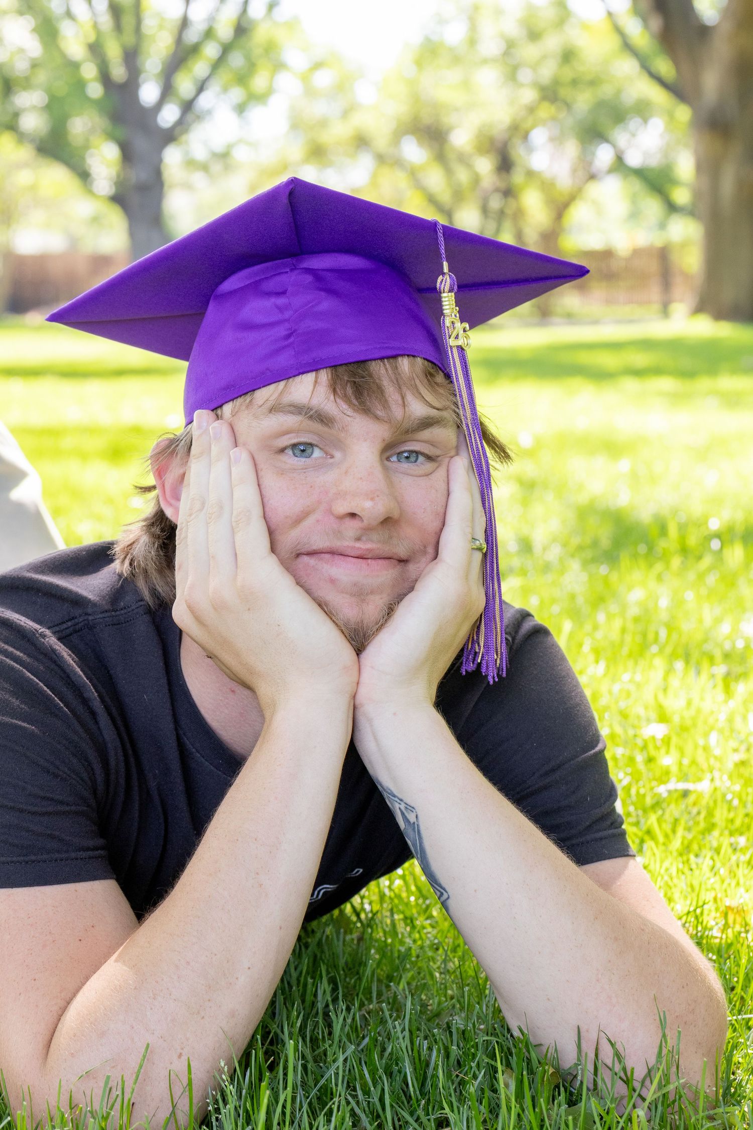 Blue-eyed young man in purple graduation cap lies on green grass resting chin on hands.