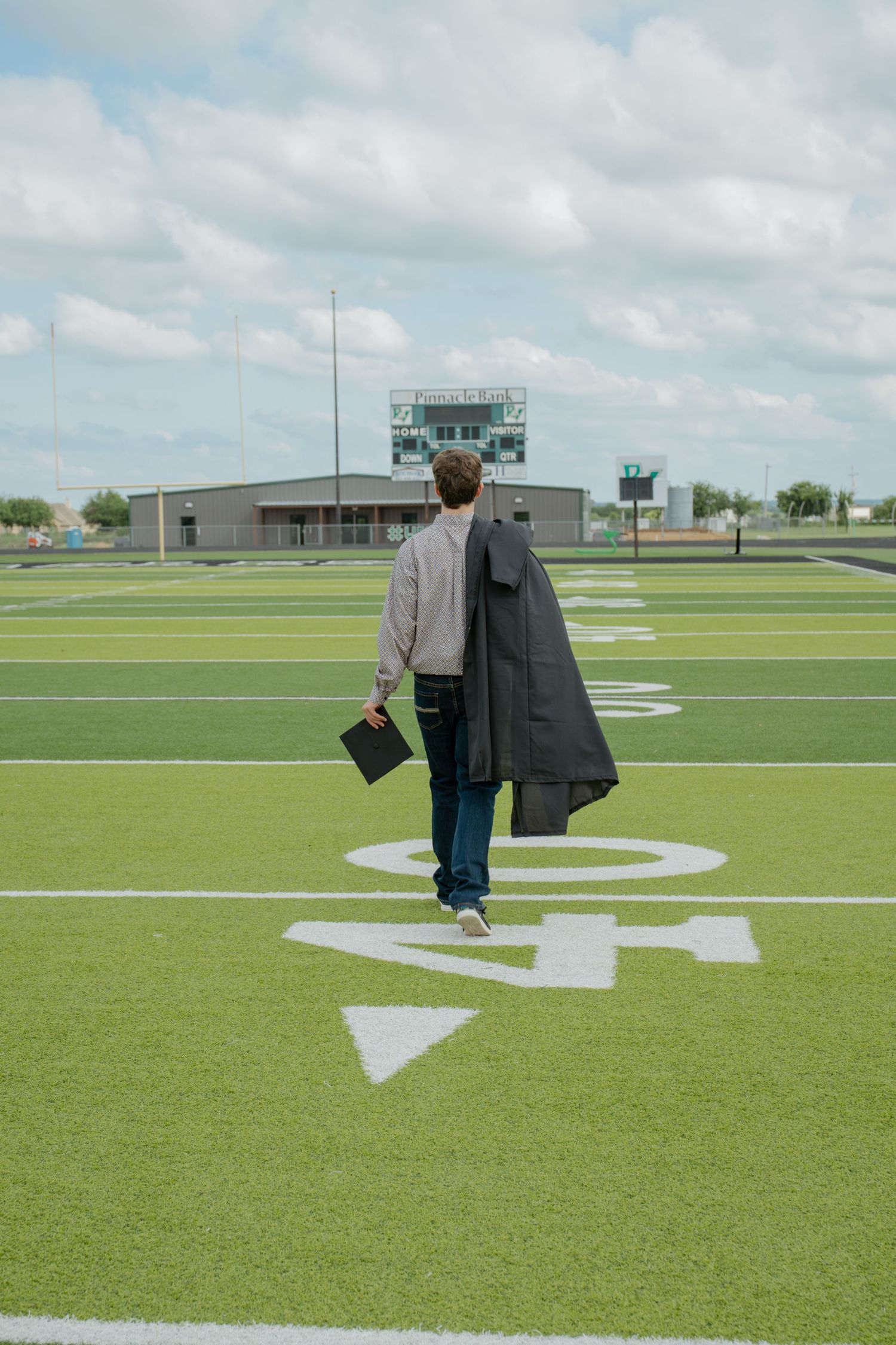 Graduate walking away on green football field carrying cap and gown under cloudy sky.