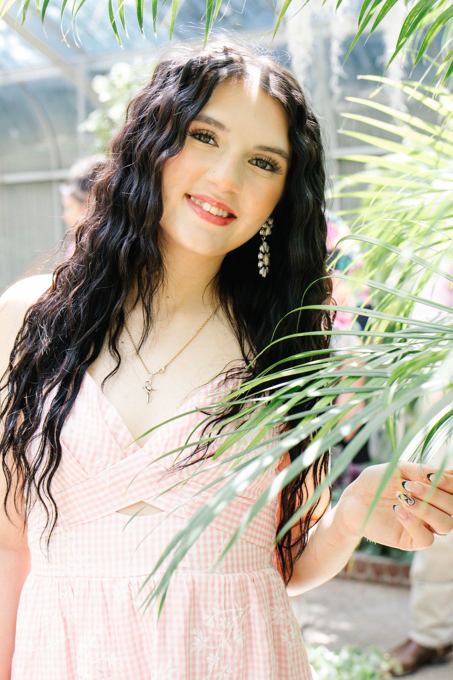 A smiling young woman with dark curly hair holds a palm leaf wearing a pink dress outdoors.