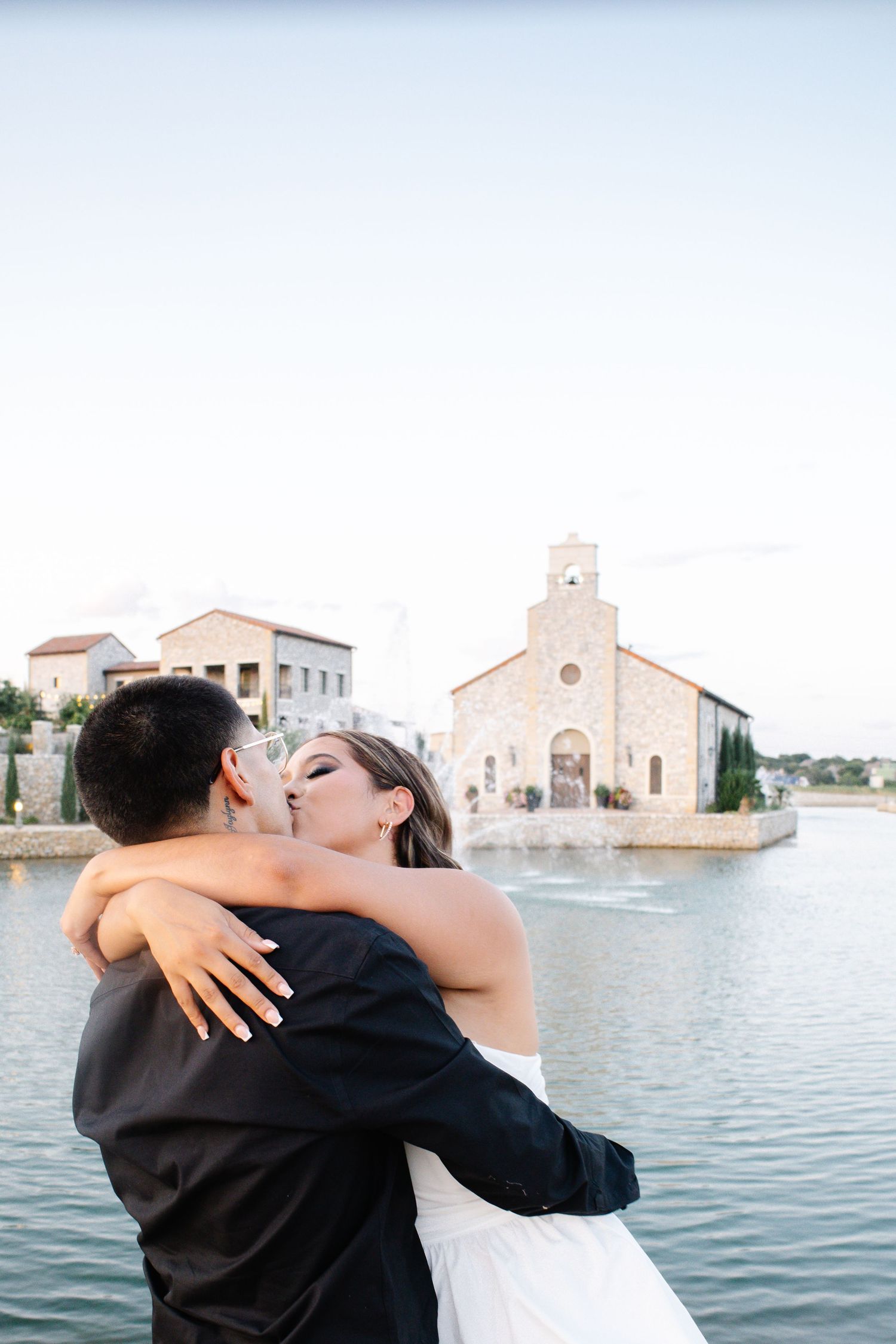 Bride and groom share a romantic kiss near a waterfront chapel during their outdoor wedding ceremony.