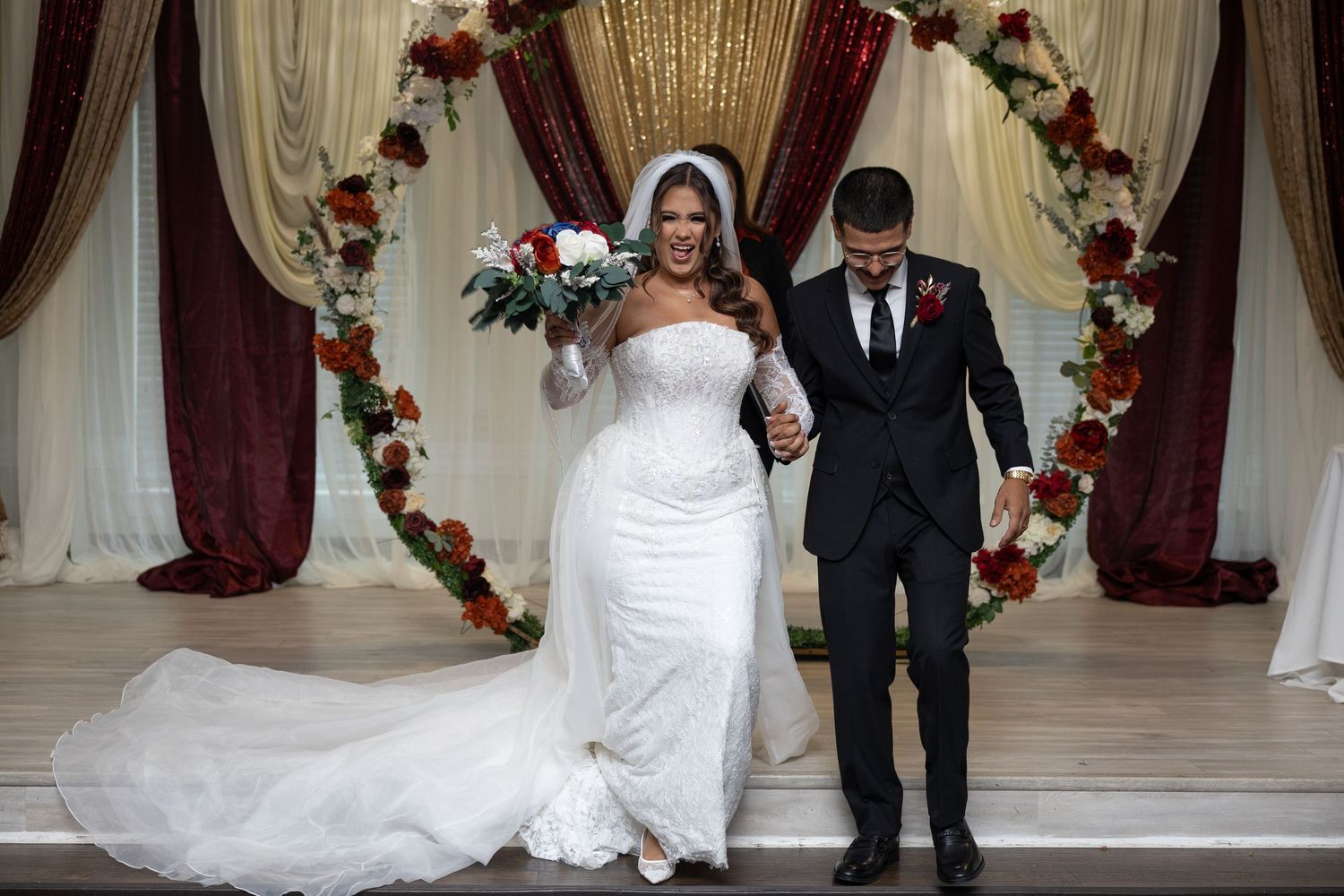 Newlyweds walk hand in hand under floral arch with red and white flowers at wedding ceremony altar.