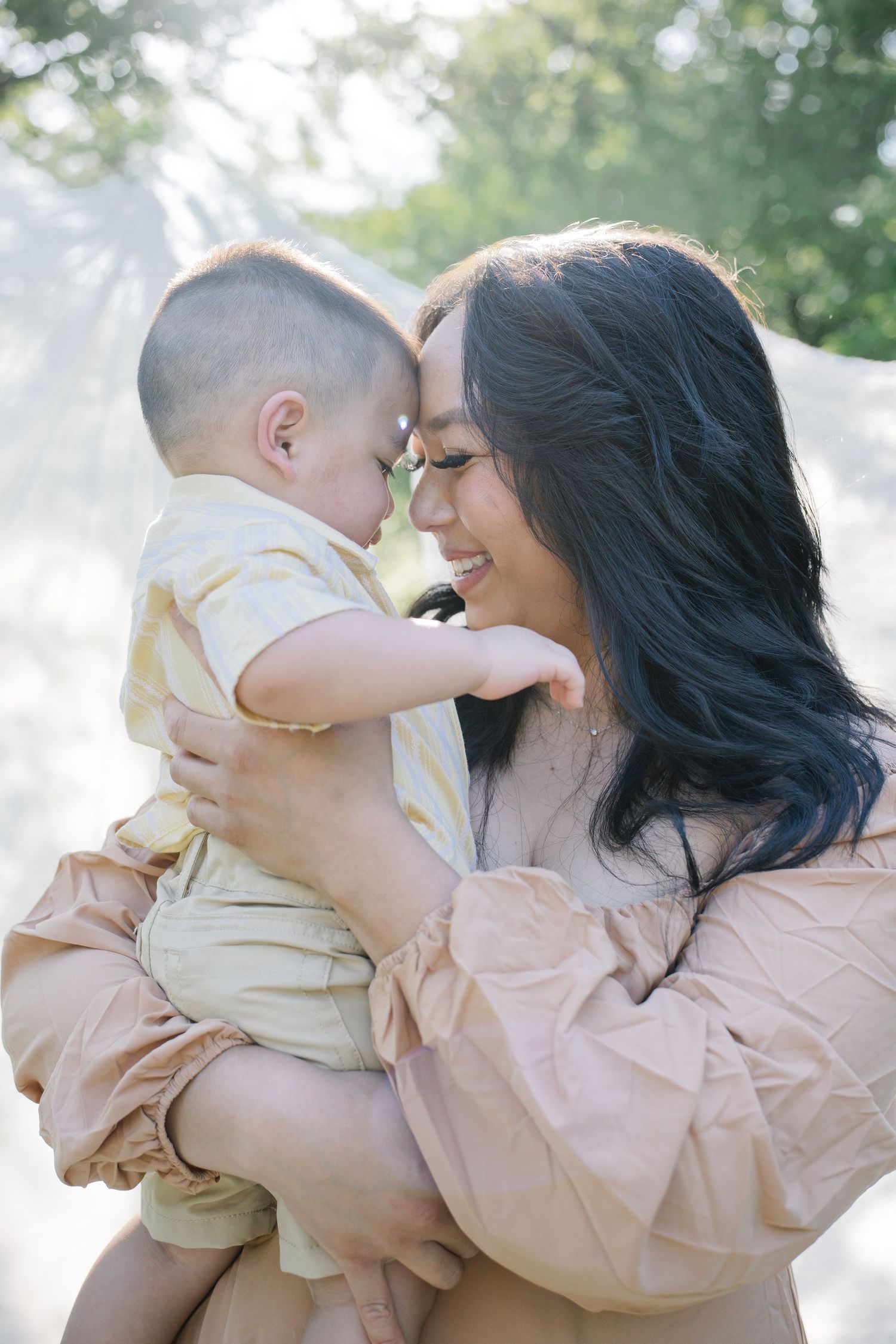 A smiling woman with dark hair holds a baby close, touching foreheads outdoors in soft natural light.