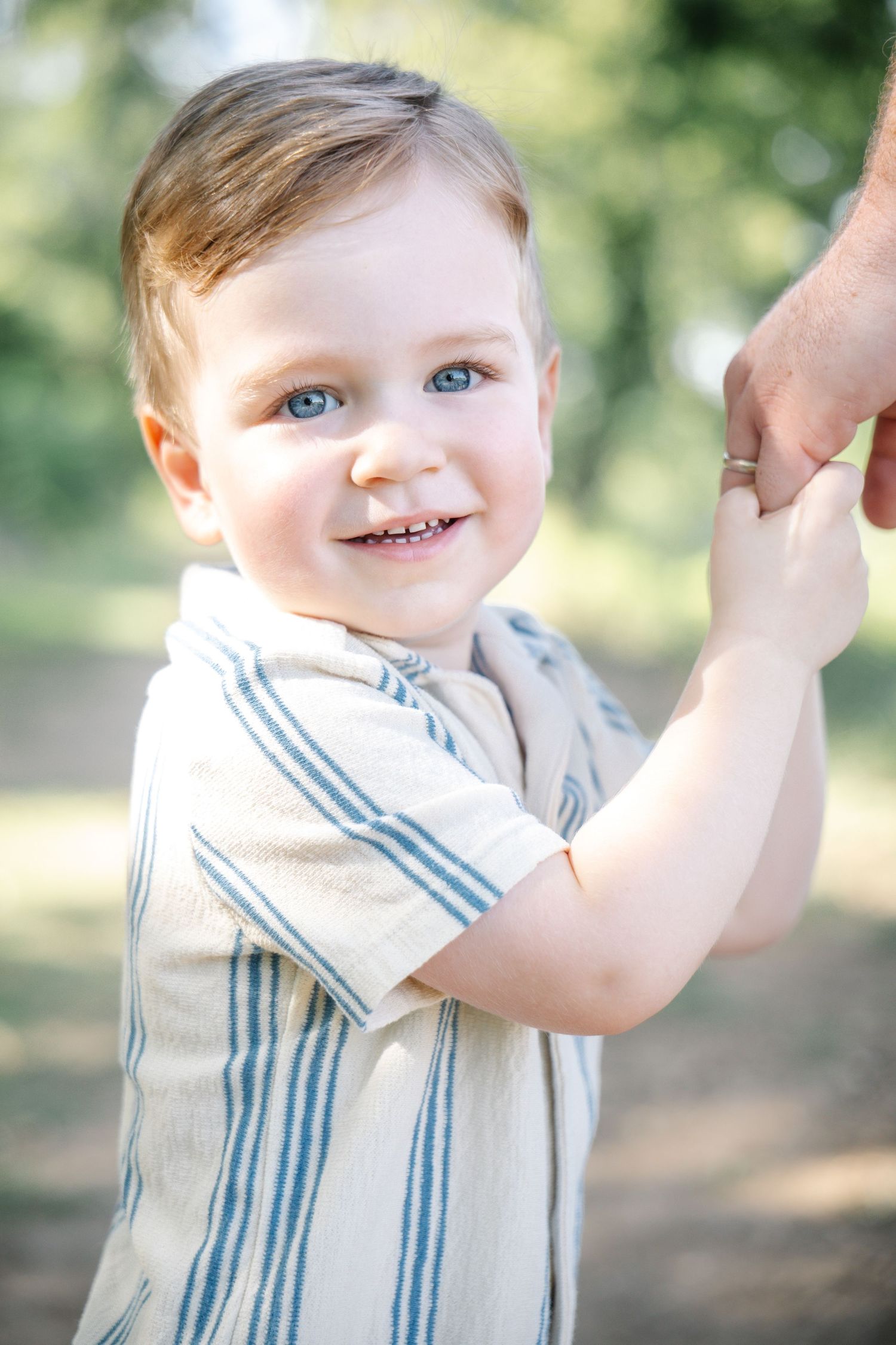 Smiling toddler boy with blue eyes holding an adult's hand outdoors in a sunny park setting.