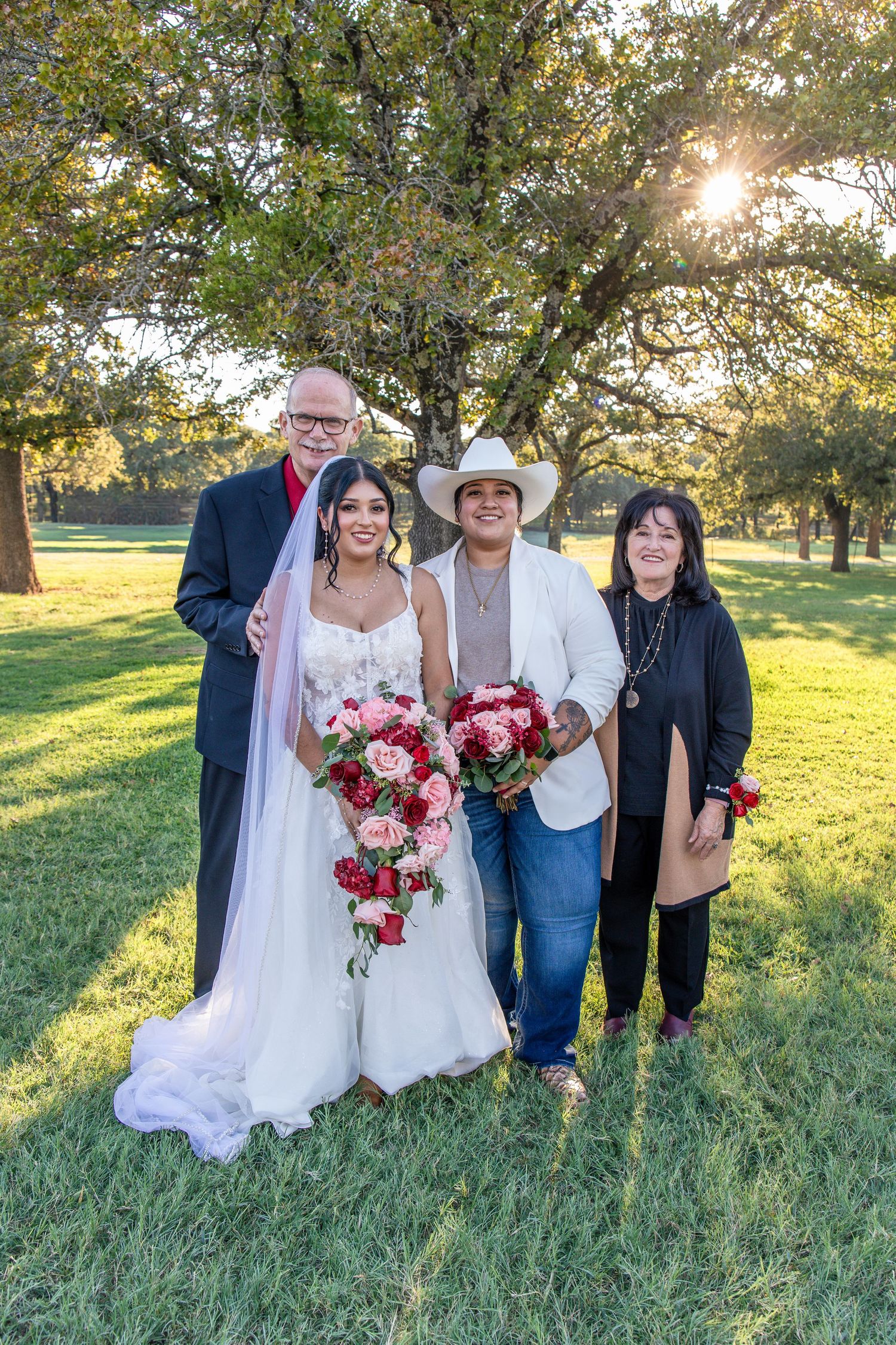 Bride in a white wedding gown and veil holding a pink and red bouquet, posing with three family members on a sunlit grassy lawn.