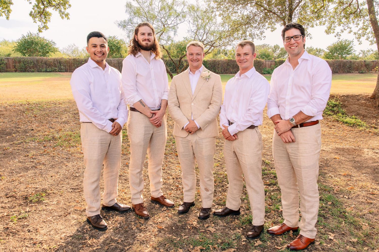 Groom and five groomsmen in white shirts and tan pants stand in a row on a rustic outdoor lawn at sunset.