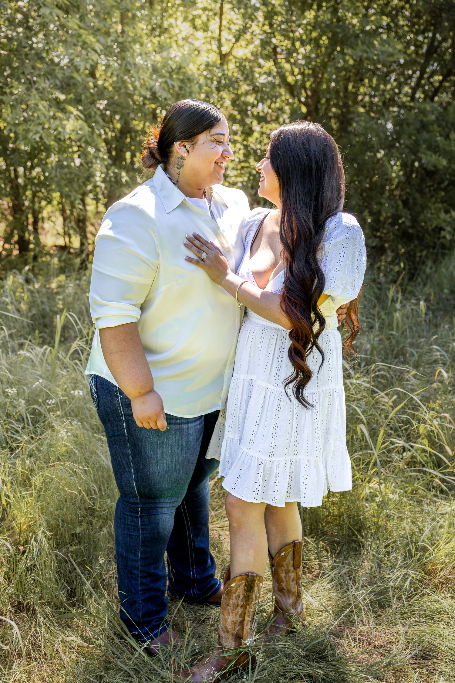 Couple smiling at each other in a lush green field, woman in white dress and cowboy boots.