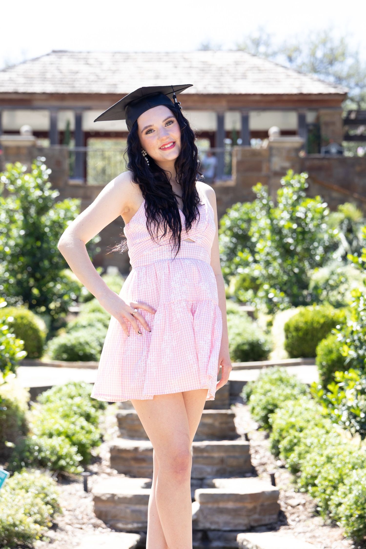 Smiling young woman in pink dress and black graduation cap posing in garden setting.