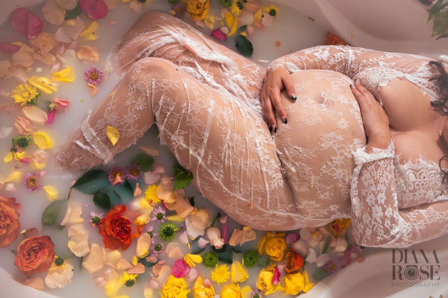 Pregnant woman in white lace reclining in a floral milk bath with colorful roses and petals.