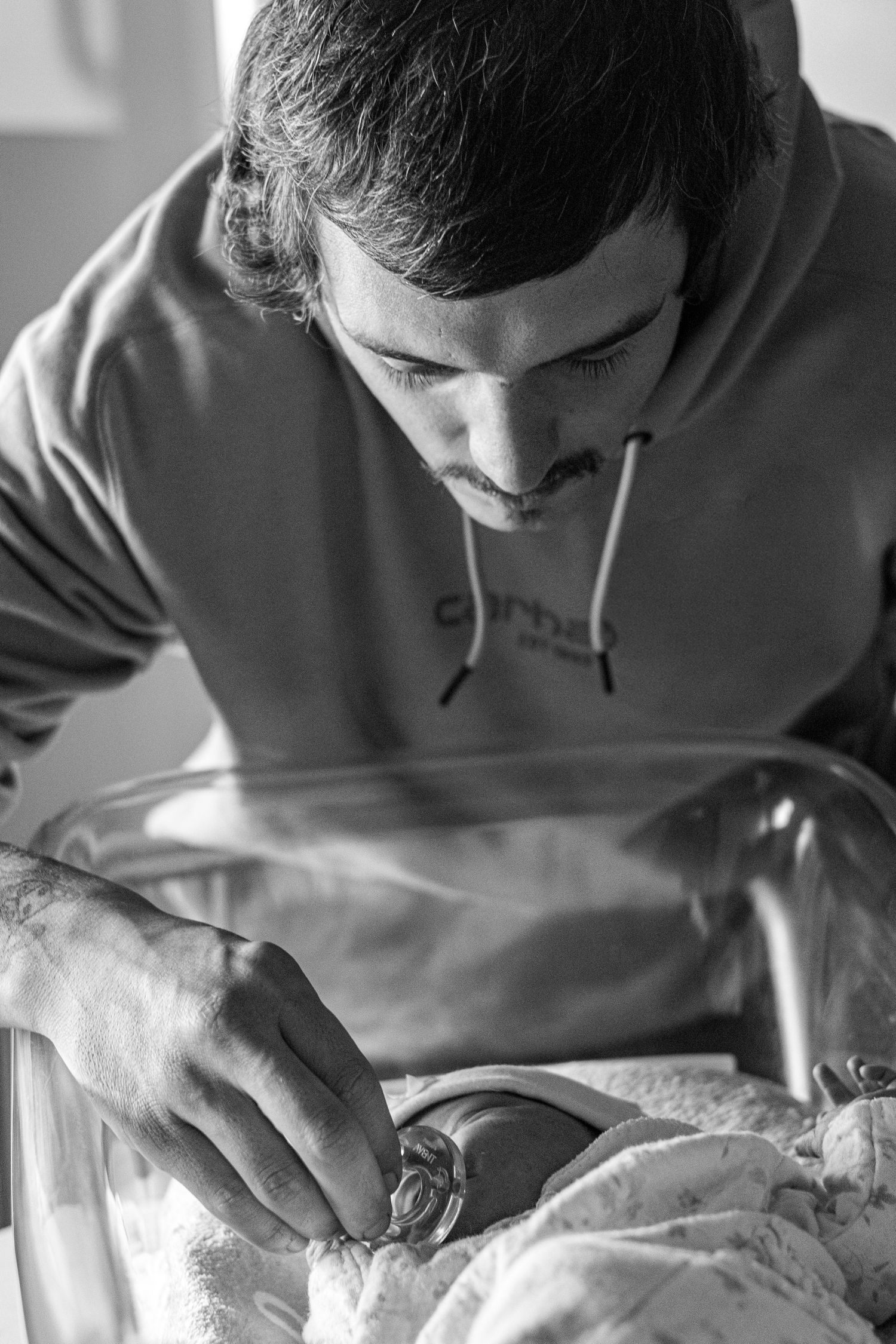 Black and white photo of a new father tenderly leaning over his newborn baby in hospital bassinet.