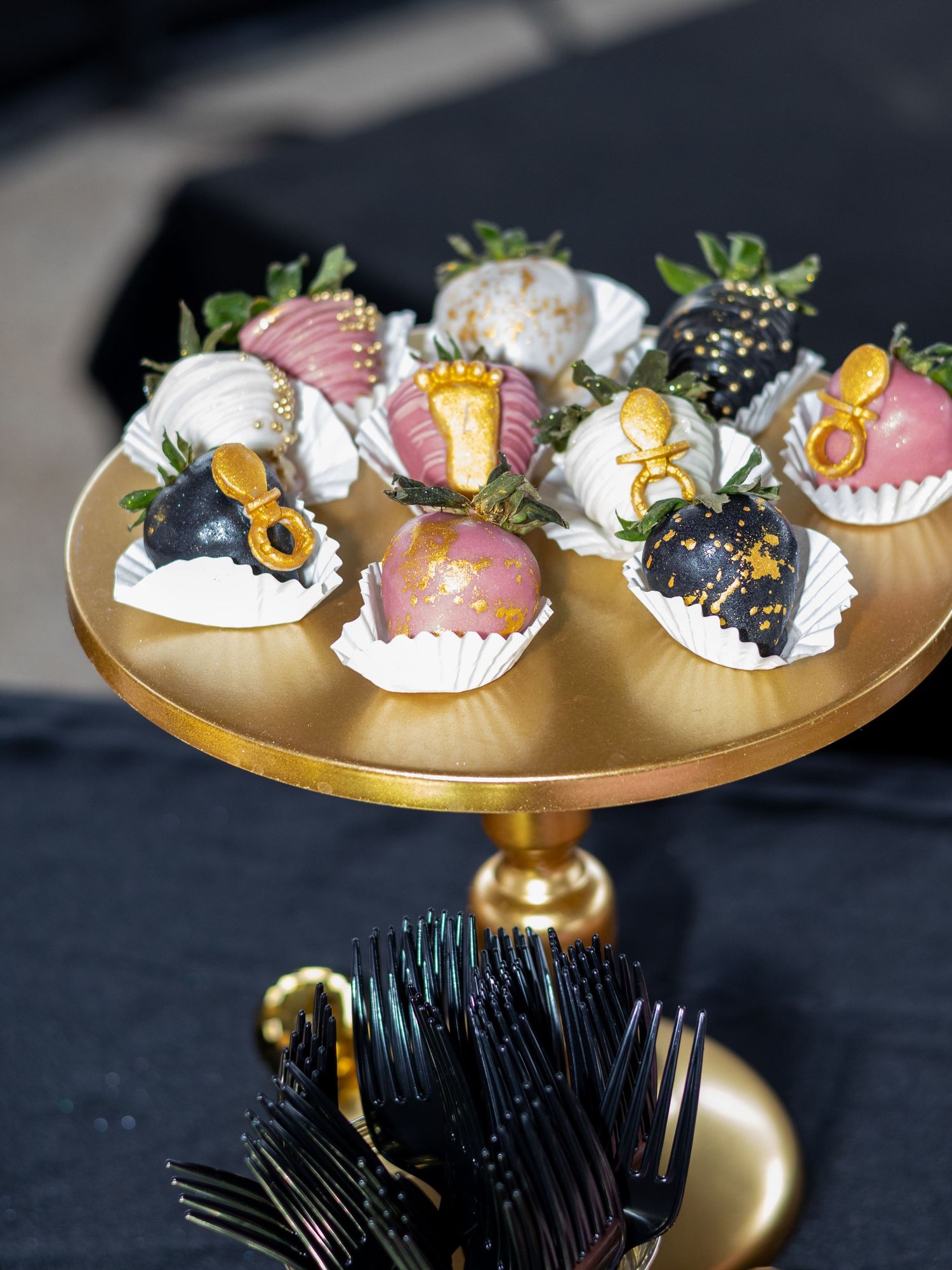 Decorated chocolate-covered strawberries with gold accents displayed on a gold cake stand with black forks.