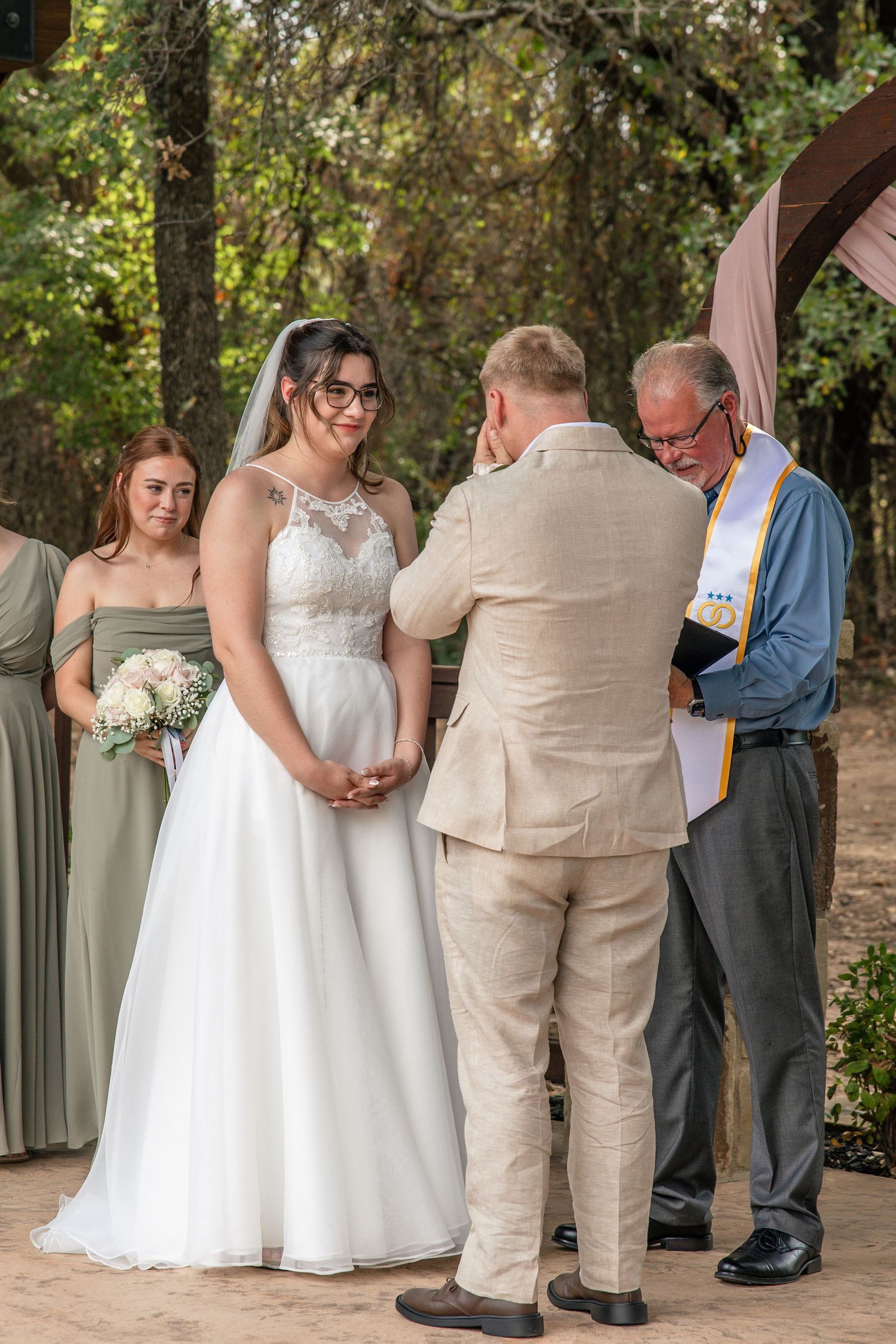 Bride in a lace wedding gown and veil faces her groom in a tan suit as an officiant leads an outdoor ceremony with a bridesmaid nearby.