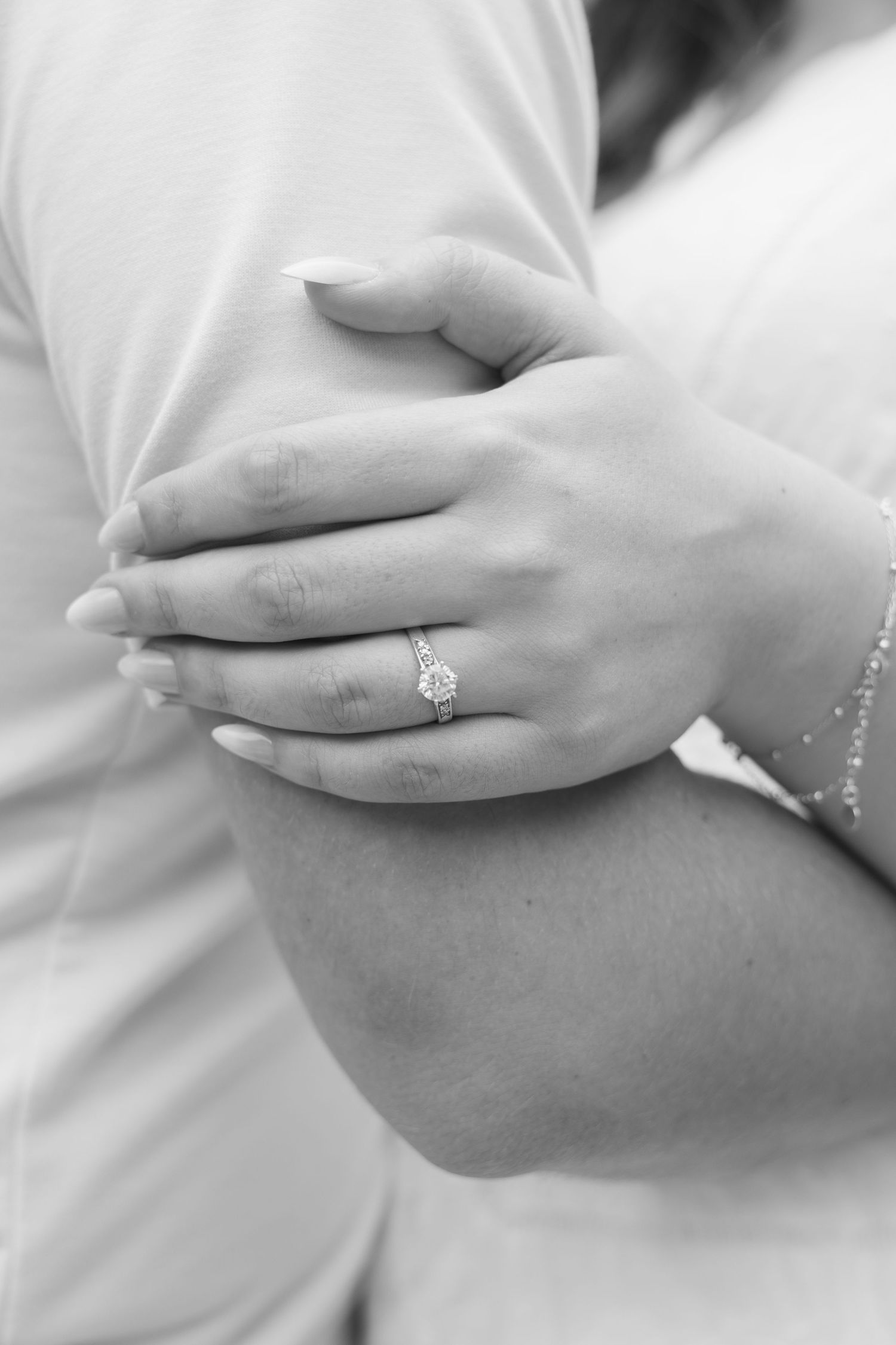 Close-up black and white photo of a woman's hand displaying a diamond engagement ring while embracing her partner.
