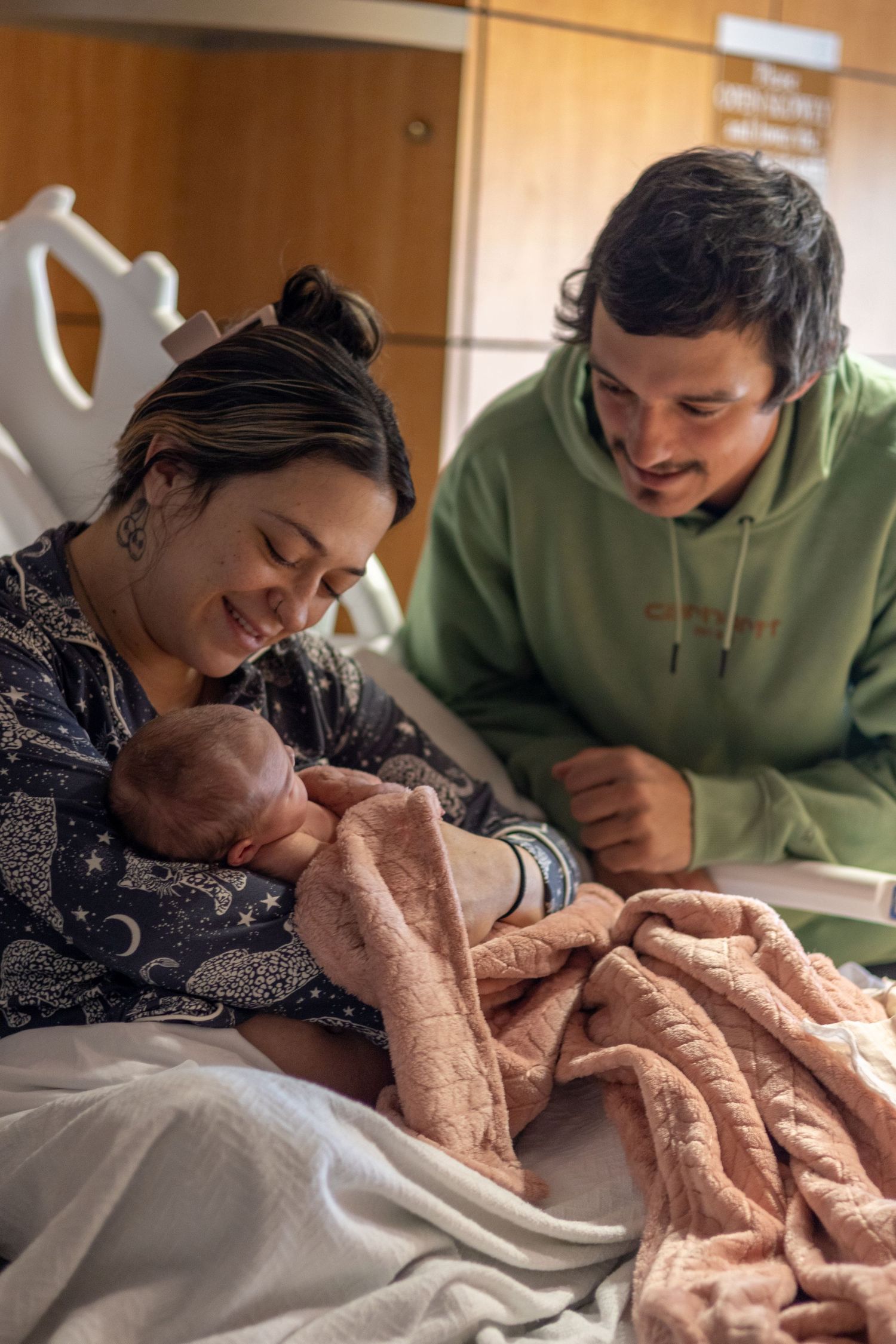 New parents smiling lovingly at their newborn baby wrapped in a pink blanket in hospital.