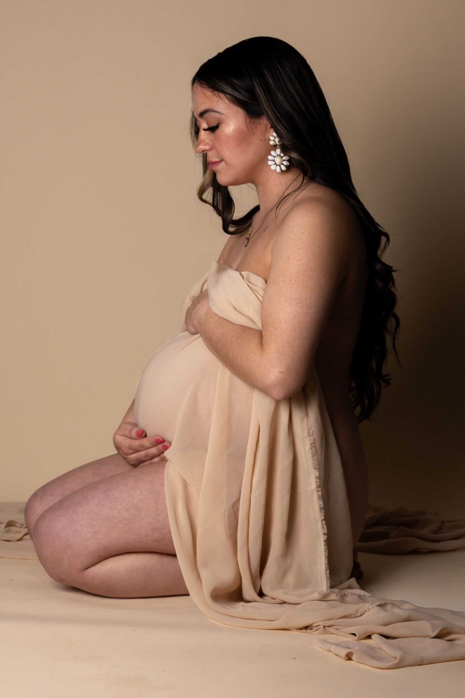 Side profile of pregnant woman in beige fabric wrap with floral earrings, holding belly.
