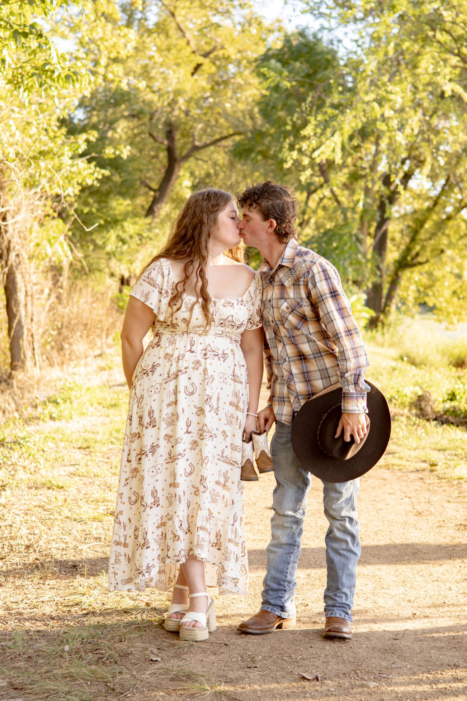 Pregnant woman and man in plaid shirt sharing a kiss on a sunlit dirt path holding baby shoes and a cowboy hat.
