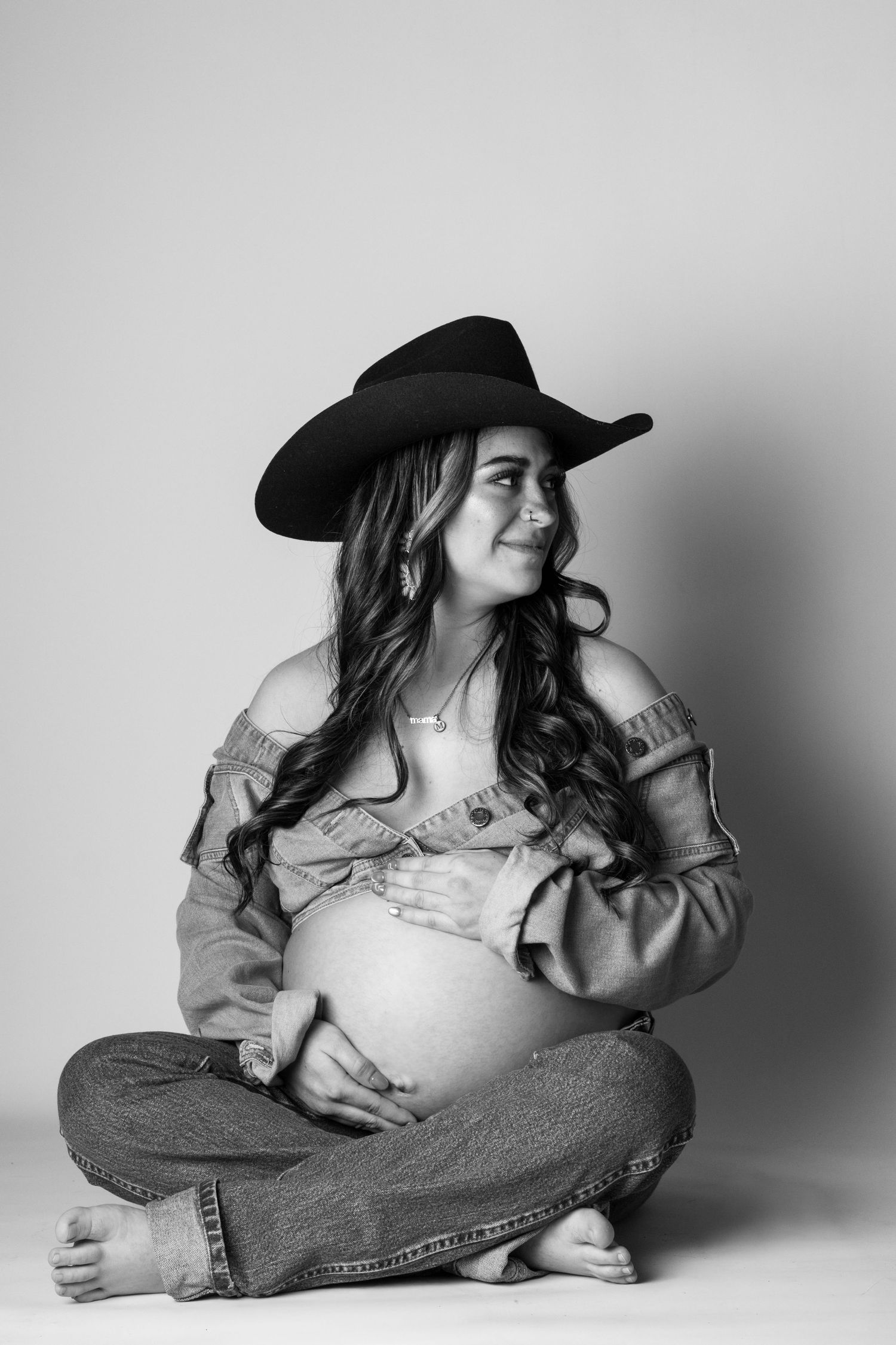 Black and white maternity photo of a pregnant woman in cowboy hat, jeans, sitting cross-legged, cradling her baby bump.